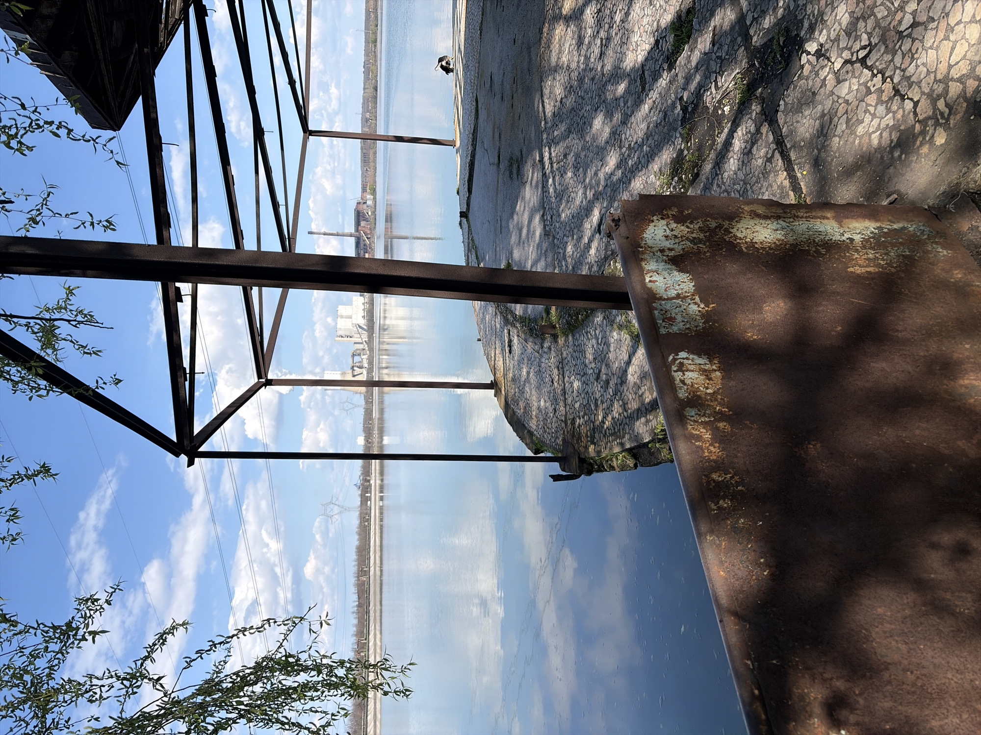 The scene is an outdoor environment in Galabovo, Bulgaria. An industrial landscape can be seen in the distance, consisting of power lines, factory buildings, and smokestacks. In the foreground, there is a weathered metal structure, with rust evident on the surface. The framework casts a shadow on the cracked and cobblestone ground. There is a body of water, possibly a river or lake, which reflects the sky and clouds. A few tree branches are visible. In the background, a bird is seen.