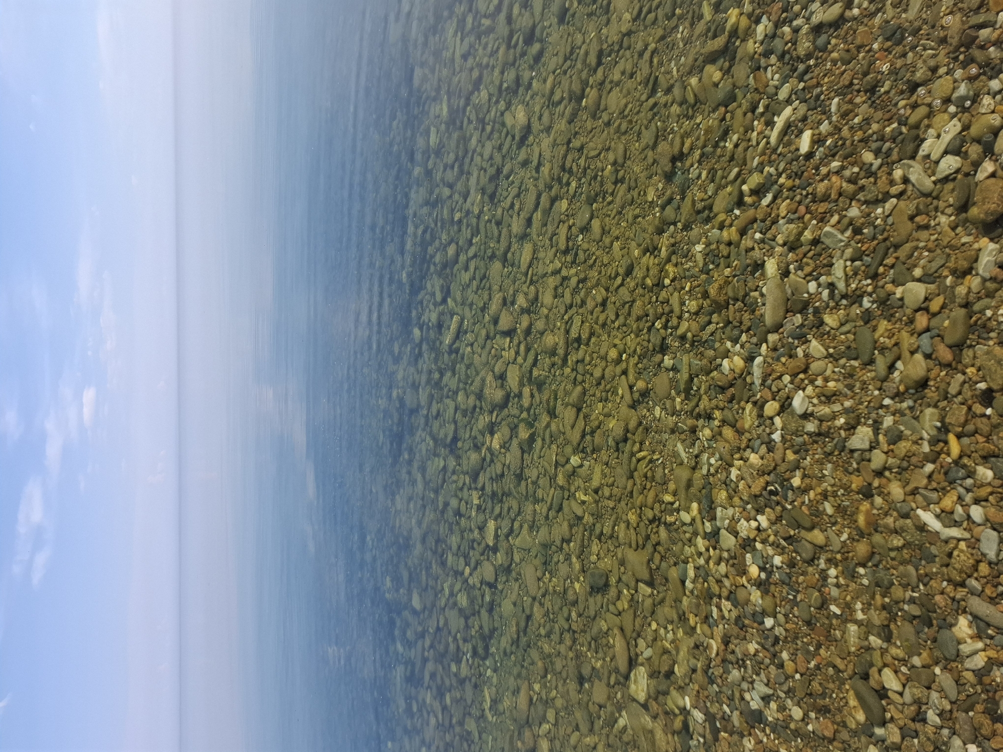 This is an outdoor, eye-level shot of a shallow body of water with a rocky bottom, with the sky visible on the left side. The water is clear, allowing the pebbles and rocks to be seen distinctly. The pebbles are of various sizes and colors, predominantly shades of brown, grey, and tan, with some lighter-colored stones interspersed. The water surface reflects the light, creating subtle patterns.
On the left side of the image, the water transitions into the sky, which is a pale blue with a few wispy white clouds. The horizon line is blurred and indistinct, suggesting a hazy atmosphere or a far-off distant view. The sunlight appears to be bright, indicating it is likely daytime. The overall scene evokes a sense of natural tranquility and the beauty of a clear, shallow water environment. There is no text visible in the image, nor are there any people or identifiable objects. The location is identified as Bugul'deika, Russia.
