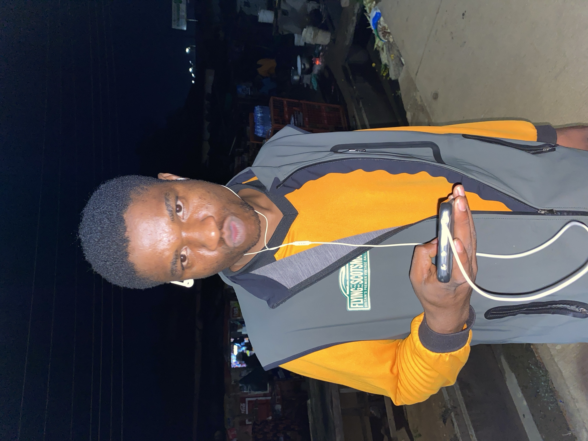 An image taken at night in Bukuru, Nigeria, features a young Black man posing for the camera. He is shown from the chest up, with short, dark, curly hair and a casual expression, his mouth slightly open. He is dressed in a vibrant orange long-sleeved shirt, layered with a grey and black vest that has zippered pockets. White wired earphones are plugged into his ears and connected to a dark-cased smartphone he holds in his right hand.
A notable detail on his vest is a green and white logo, which reads "FLYING SCOTSMAN" with the phrase "BRITAIN'S PREMIER VINTAGE RALLY" beneath it.
The background, dimly lit by artificial sources, suggests an outdoor, urban or semi-urban street scene, possibly a market area or stalls. Various indistinct objects like crates, bottles of water, and some refuse can be faintly seen, indicating a bustling environment even at night. The overall atmosphere is nocturnal and candid.