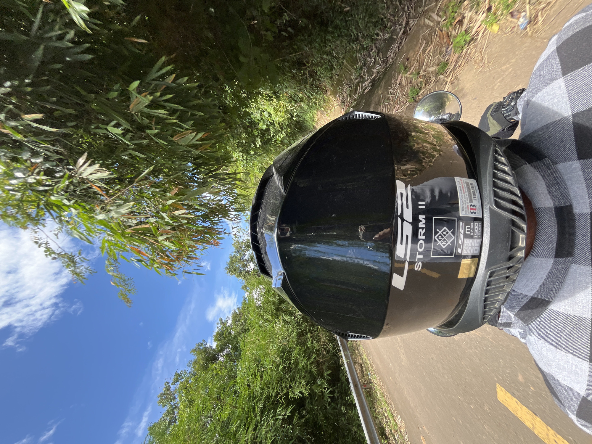 In a first-person perspective, a black LS2 Storm II motorcycle helmet is the central focus of the image. The helmet is adorned with stickers, including one with the text "TEM CHONG HANG GIA" and another displaying certification marks like "ECE 22.06" and "1500 g". The rider, wearing a gray and white plaid shirt, is on a motorcycle. They are traveling on a paved road with a yellow line marking, bordered by lush green foliage and bamboo trees. The scene appears to be in a rural or natural setting, likely Son Dien, Vietnam, given the context. The sky is a vibrant blue with scattered white clouds, suggesting good weather. The road curves gently to the right, disappearing into the verdant landscape.