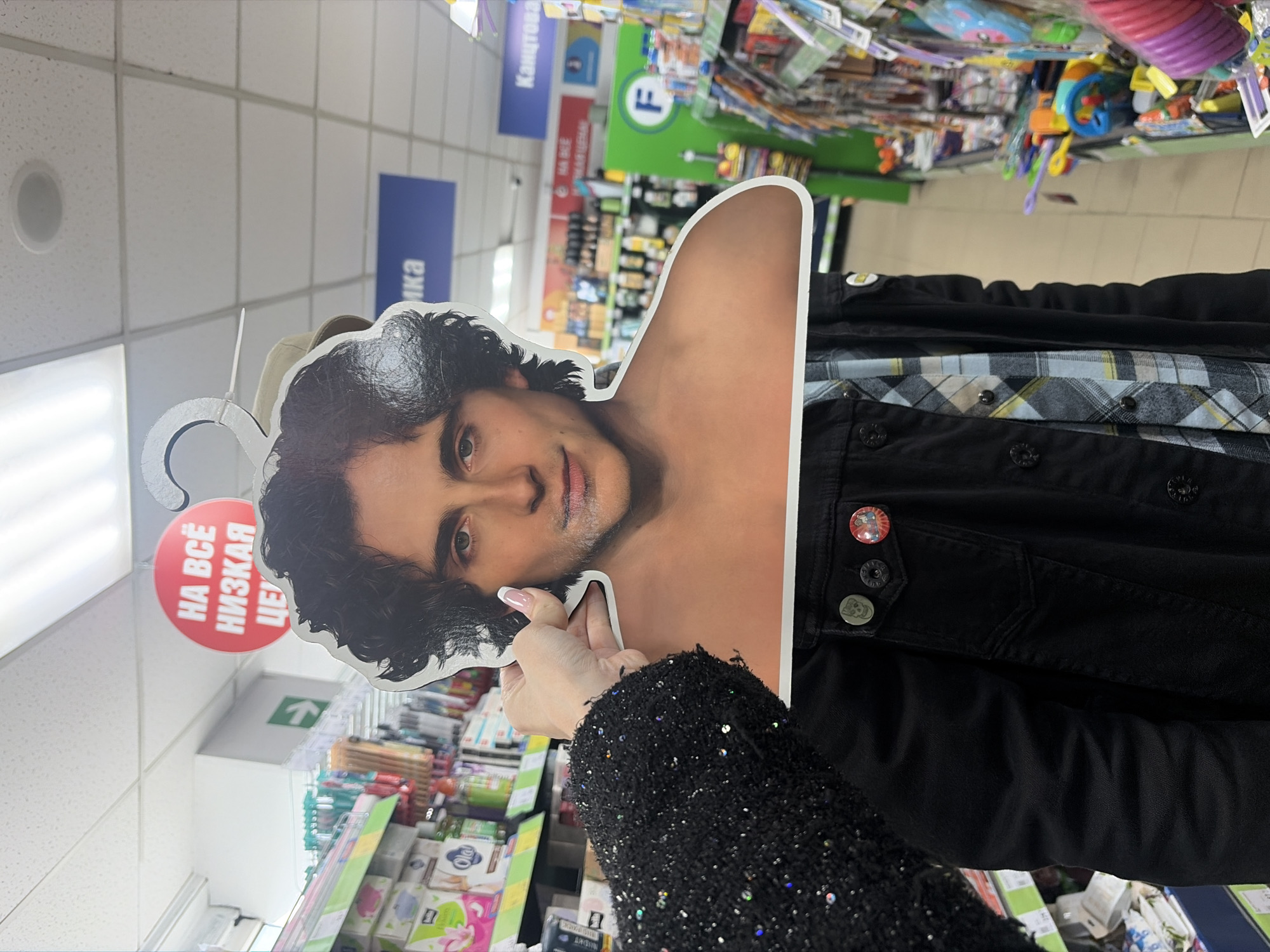 A person in a black sequined garment is holding a cardboard cutout of a man's head and bare shoulder in a retail store. The person is facing the camera, but only their arm is visible. The cutout is predominantly white and features a dark-haired man with light skin. The background consists of shelves stocked with various products, suggesting a grocery or department store. A sign in Russian with a red circle is also visible, partially obscured by the cutout. Another sign is visible with the letter 'F' on it. Based on the Russian language sign, it is possible that the location is in Saint Petersburg, Russia.