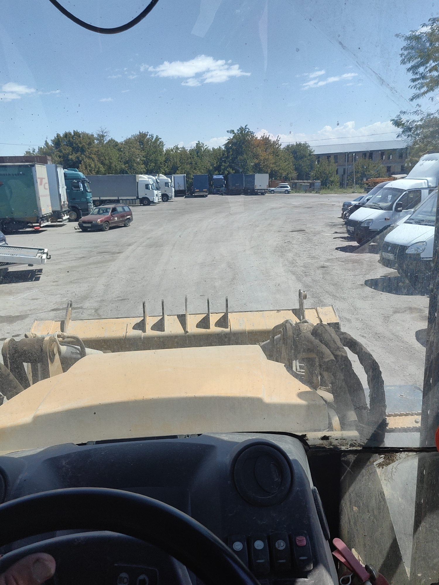 The image, taken from the perspective of a heavy machinery operator, shows a parking lot with several trucks and cars under a clear sky with scattered clouds. In the foreground, a yellow loader's bucket and frame are visible, suggesting the operator is in a construction or industrial setting. Several semi-trailer trucks are parked in rows, some with white cabs and others with dark blue trailers. A red station wagon is parked in the middle of the lot. In the background, trees and a large, multi-story building with a light-colored facade are visible. The overall environment appears to be an outdoor commercial or industrial area. The specific location of Shymkent, Kazakhstan is not discernible from the visual elements within the image itself.