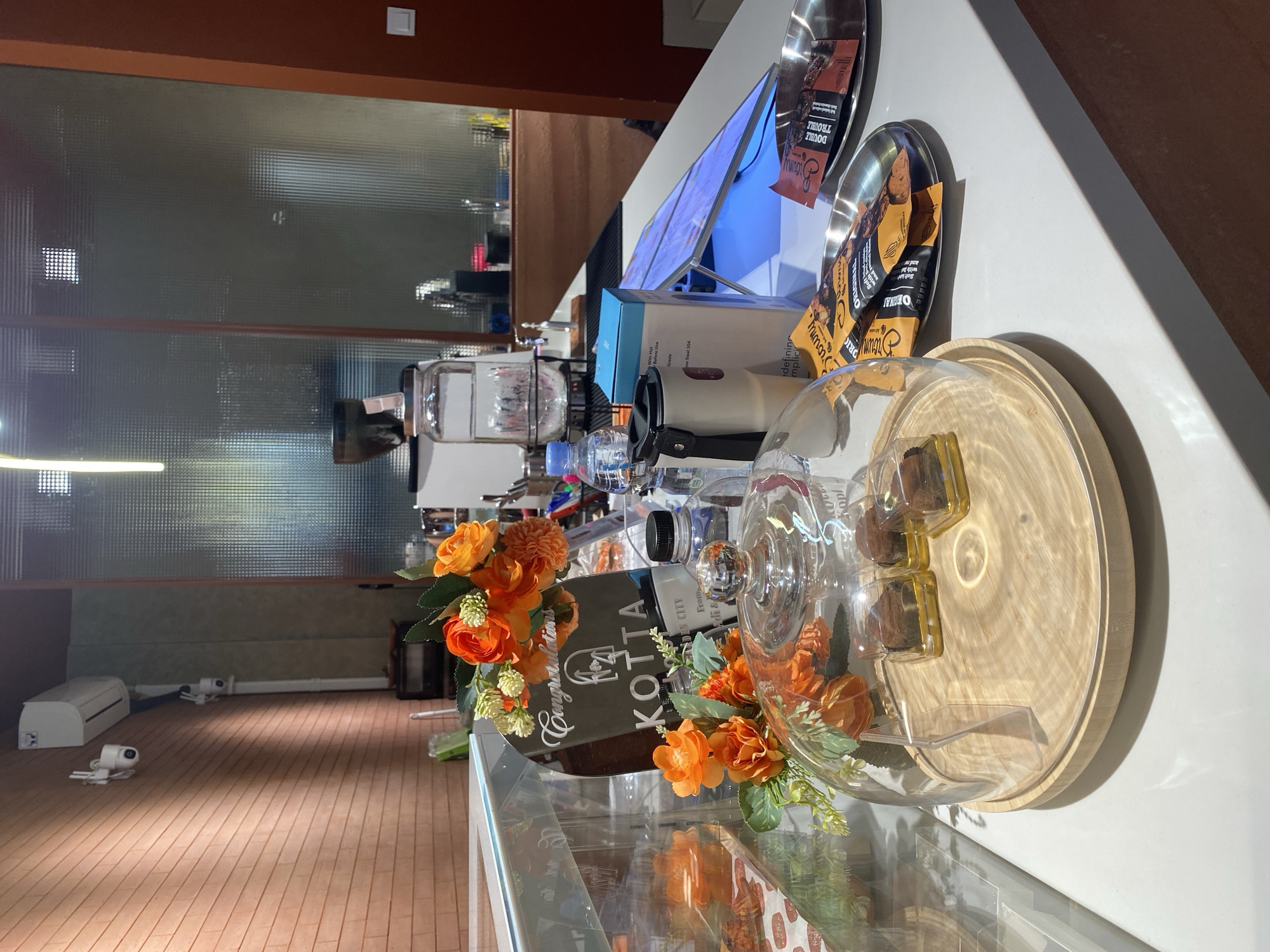 The image captures a counter area within what appears to be a cafe or bakery in East Jakarta, Indonesia. In the foreground, a glass dome covers a wooden tray holding four individually packaged chocolate brownies. To the left, artificial orange flowers with green foliage are arranged, extending over the display. Behind the flowers, a mirrored sign with "KOTTA" and the text "Congratulations" is visible.
On the counter, to the right of the brownies, are several items: a metallic tray holding two small packages labeled "Sikuning Brownies" (one "Original" and one "Double Trouble"), a white ceramic mug with a black lid, and a blue and white box. Further back, a large glass dispenser filled with water sits on a stand, and various bottles, including a blue plastic water bottle, are present. In the background, a glass partition with a textured pattern separates the counter area from what seems to be the back of the establishment, where kitchen equipment and shelving are dimly visible. On the wall to the far left, an air conditioning unit and security cameras are mounted. The lighting is artificial, creating a warm ambiance. There are no people visible in the scene.
