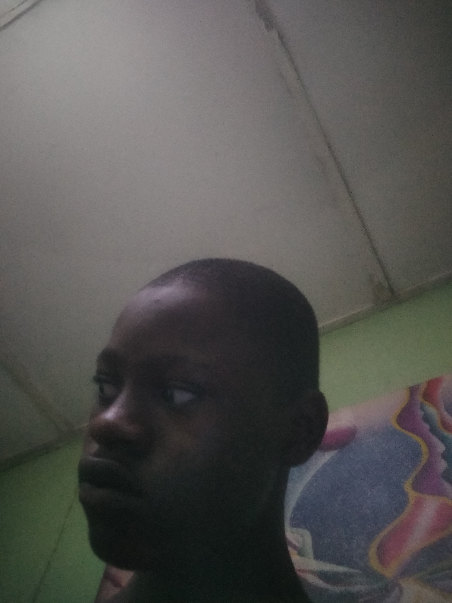 A close-up, dimly lit image captures a young person, likely a child or teenager, with dark skin and short, dark hair. The individual is positioned slightly off-center, facing to their left in the frame, with their head tilted slightly upwards. Their eyes are cast towards the right side of the image (viewer's left), suggesting a sideways glance or a thoughtful gaze. The lighting is low, creating shadows on the right side of their face and accentuating their facial features on the left.
The background reveals an indoor setting. Above the person's head, a pale, possibly white or light gray, ceiling is visible, showing some lines, seams, or minor discoloration. To the left and lower right of the frame, a plain, light green wall is visible. Behind the person's right shoulder, a colorful, abstractly patterned object, likely a pillow or blanket, features swirling shapes in shades of blue, purple, yellow, and pink. No specific activity is taking place other than the person's attentive gaze. There is no visible text in the image. The overall impression is one of quiet contemplation in a simply furnished, low-light room.