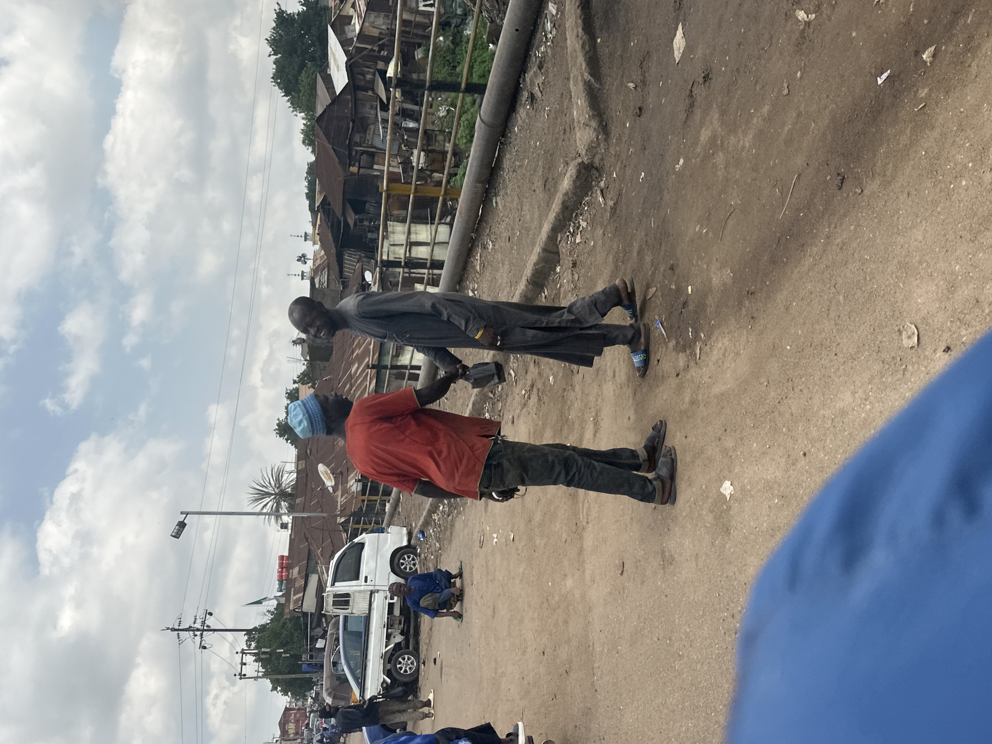 The image depicts a street scene in Jos, Nigeria. Two men are the primary subjects. The man in the foreground is wearing a red t-shirt and dark pants, with a light blue cap on his head. He appears to be holding something in his hand. Behind him, another man is dressed in a dark grey robe and pants. They are standing on a dusty, unpaved road.
In the background, a variety of structures can be seen, including buildings with corrugated metal roofs, suggesting a densely populated urban or semi-urban area. There are also vehicles present, including a white truck and what appears to be a smaller van or car. In the mid-ground, a person is seen crouching near the white truck.
The sky is partially cloudy, indicating it is daytime. The overall impression is a busy, everyday street scene in Nigeria, possibly in a market or residential area. The image seems to be taken at eye-level, from someone walking or standing on the street. No visible text is present in the image.
