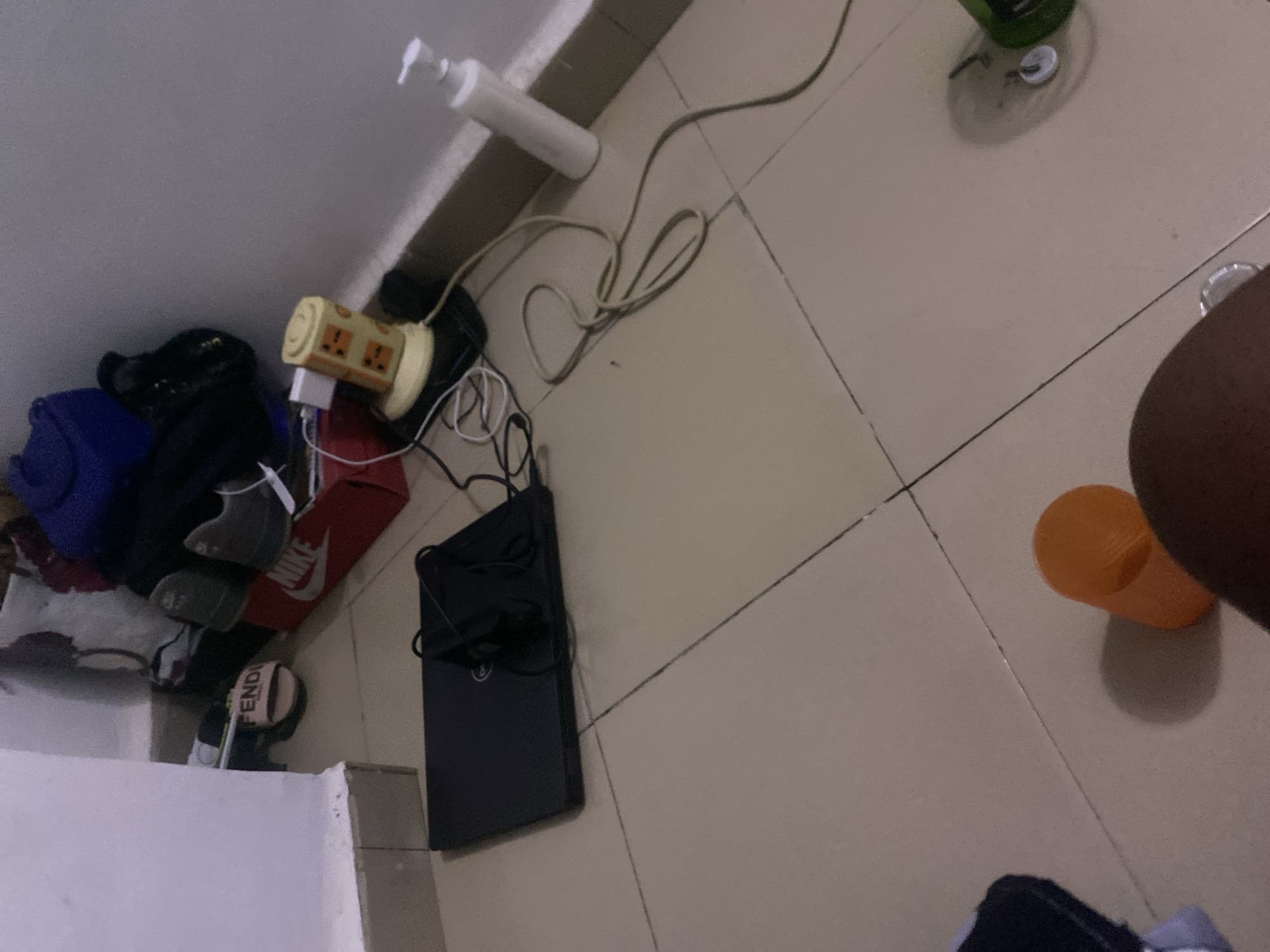 The image depicts a floor scene in a room, likely a bedroom or living area, with beige tiled flooring. In the foreground, a person's leg and thigh are visible, casting a shadow. To the left of the leg, an orange plastic cup is placed on the floor. A dark-colored laptop with a Dell logo is positioned in the middle of the frame, with a pair of black earphones on top. To the right of the laptop, a yellow multi-plug adapter with several sockets is plugged into a white power adapter. Various electrical cords are tangled around the adapter and extend across the floor. Further to the right, a white pump bottle, possibly containing soap or lotion, stands upright. In the background, there are several items including a red Nike shoebox, a pair of gray shoes, and some bags. The wall in the background is light-colored. The overall scene suggests a casual, somewhat cluttered personal space. The specific location in Howayi, Nigeria, cannot be determined from the visual information provided.