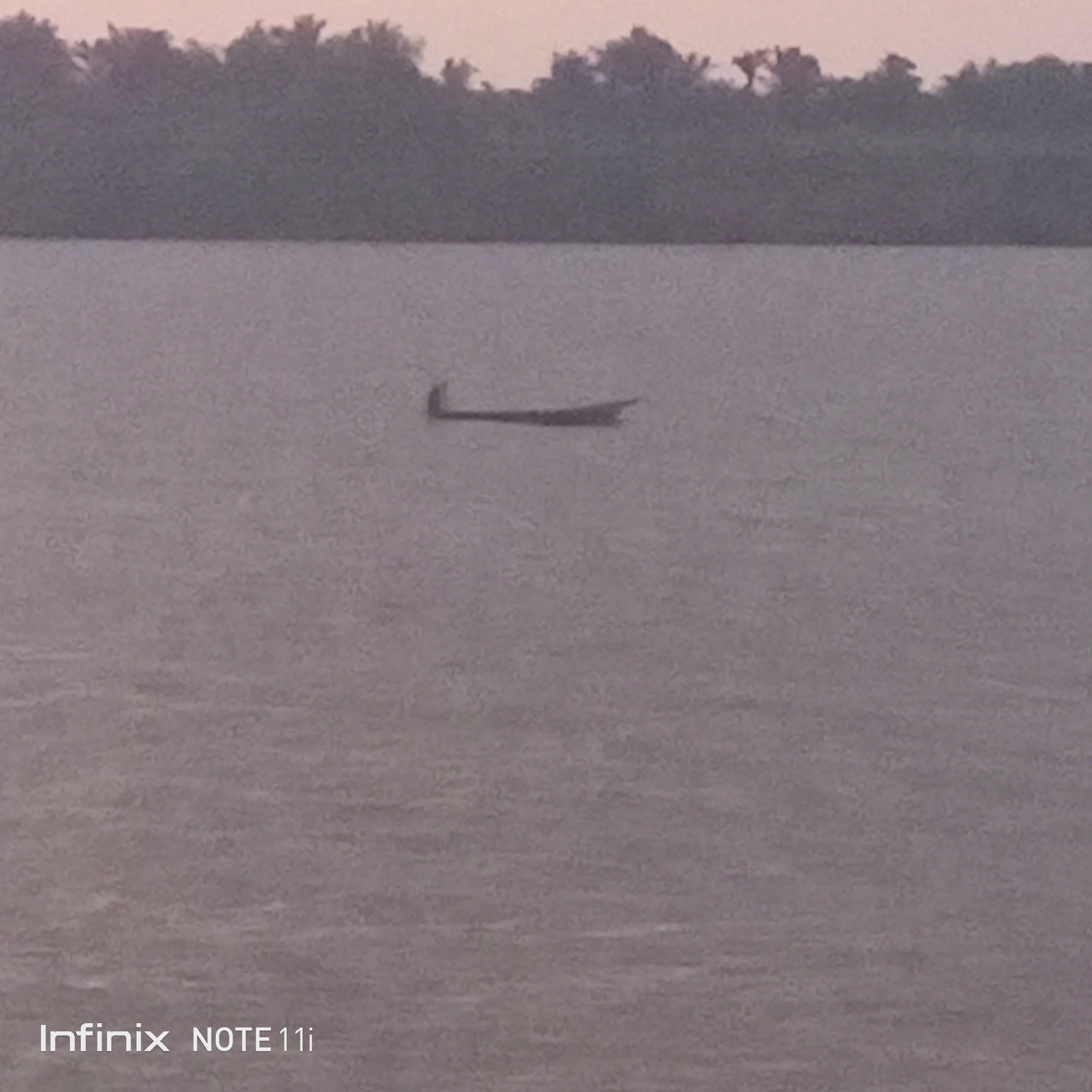 The photograph depicts a flat body of water, presumably a river or large lake, reflecting a muted, overcast sky. In the middle distance, a small wooden boat is visible, with a person in the boat, likely navigating the water. The shoreline, densely vegetated with trees, is faintly discernible in the background under a sky that hints at either dawn or dusk. The photograph was taken in Atani, Nigeria.