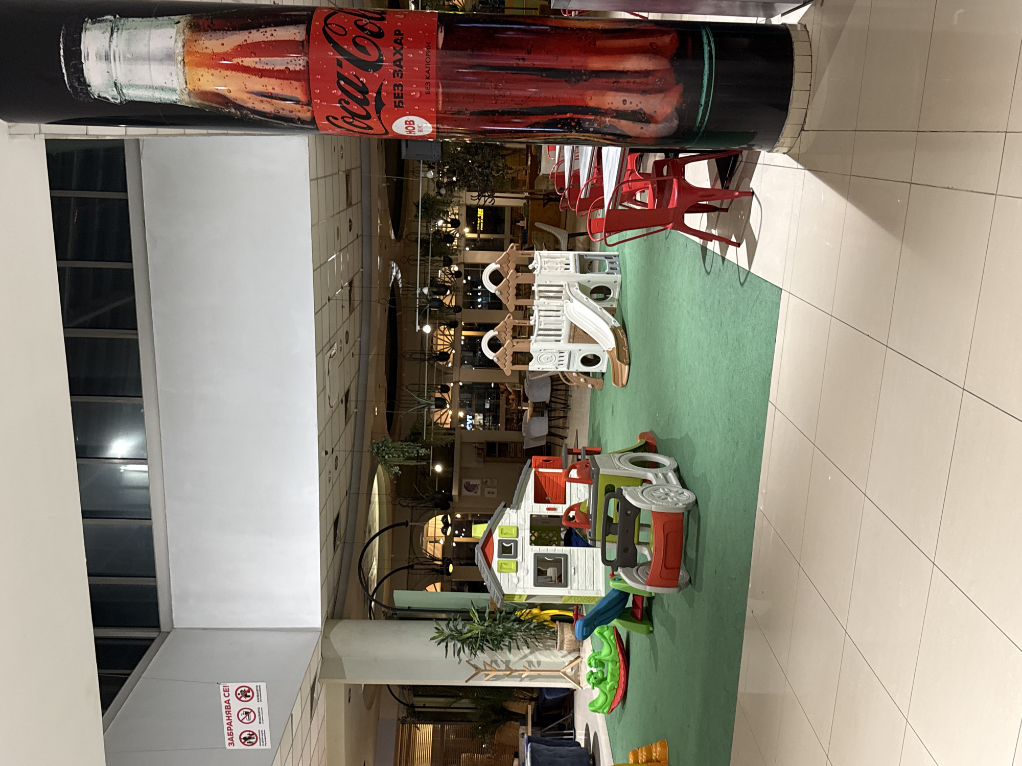 The image depicts an indoor playground located in an unidentified city. The area features various play structures, including a multi-colored playhouse with a slide and a two-story white play structure with a slide. The play area is situated on a green carpet. The background suggests a dining area with tables and red chairs. Above, there's a large Coca-Cola advertisement. Signage in a non-English language is visible on a pillar. The overall environment appears to be within a shopping mall or a similar commercial complex. There are no people visible in the immediate playground area.
