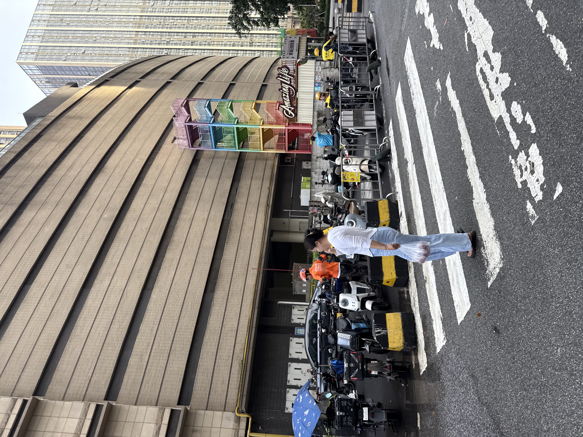 This is a full, eye-level, outdoor shot of a street scene in Haizhu District, China. A woman, wearing a white shirt and light blue pants, is walking across a crosswalk. She is holding a plastic bag containing red objects. In the background, a row of parked electric scooters and a black car are visible. Behind the vehicles, a large, curved, beige building with horizontal paneling dominates the scene. A colorful, rainbow-themed outdoor staircase is attached to the side of the building. Further in the background, a modern, tall glass building rises into the sky. The ground is wet, suggesting recent rain. Several delivery drivers in yellow and orange uniforms are standing near their scooters. The overall atmosphere appears to be a busy urban commercial area.
