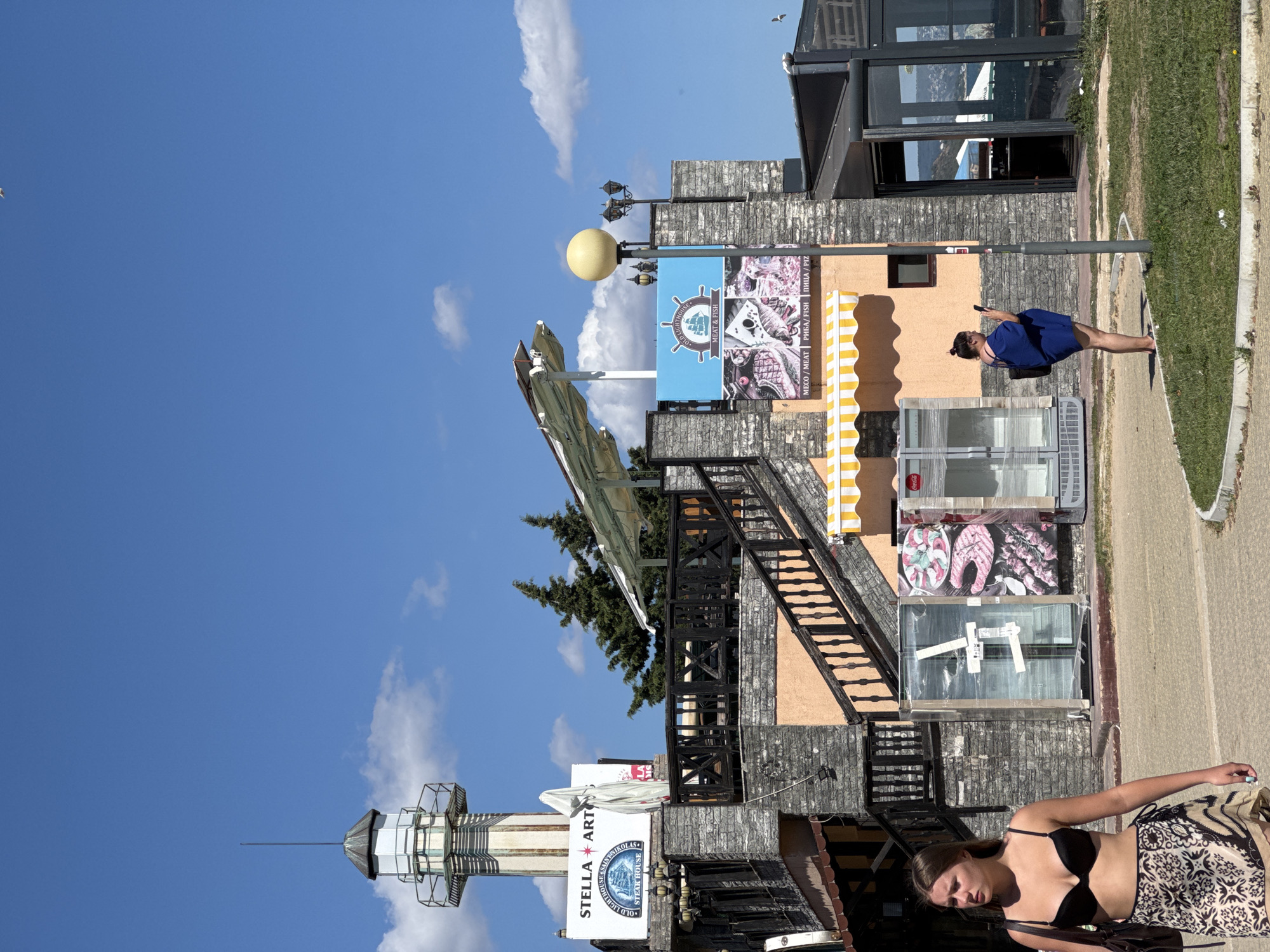The image depicts a sunny day in Varna, Bulgaria, with a clear blue sky and scattered clouds. In the background, a lighthouse-like structure stands tall, adorned with signage that reads "STELLA ART" and "OLD LIGHTHOUSE STEAK HOUSE." Adjacent to it is a restaurant with outdoor seating and a bright yellow awning. A woman in a blue dress stands near a lamppost, looking at her phone. In the foreground, another woman walks by, wearing a black bikini top and a patterned skirt. The area appears to be a promenade or walkway along the coast, with glimpses of the sea in the distance.