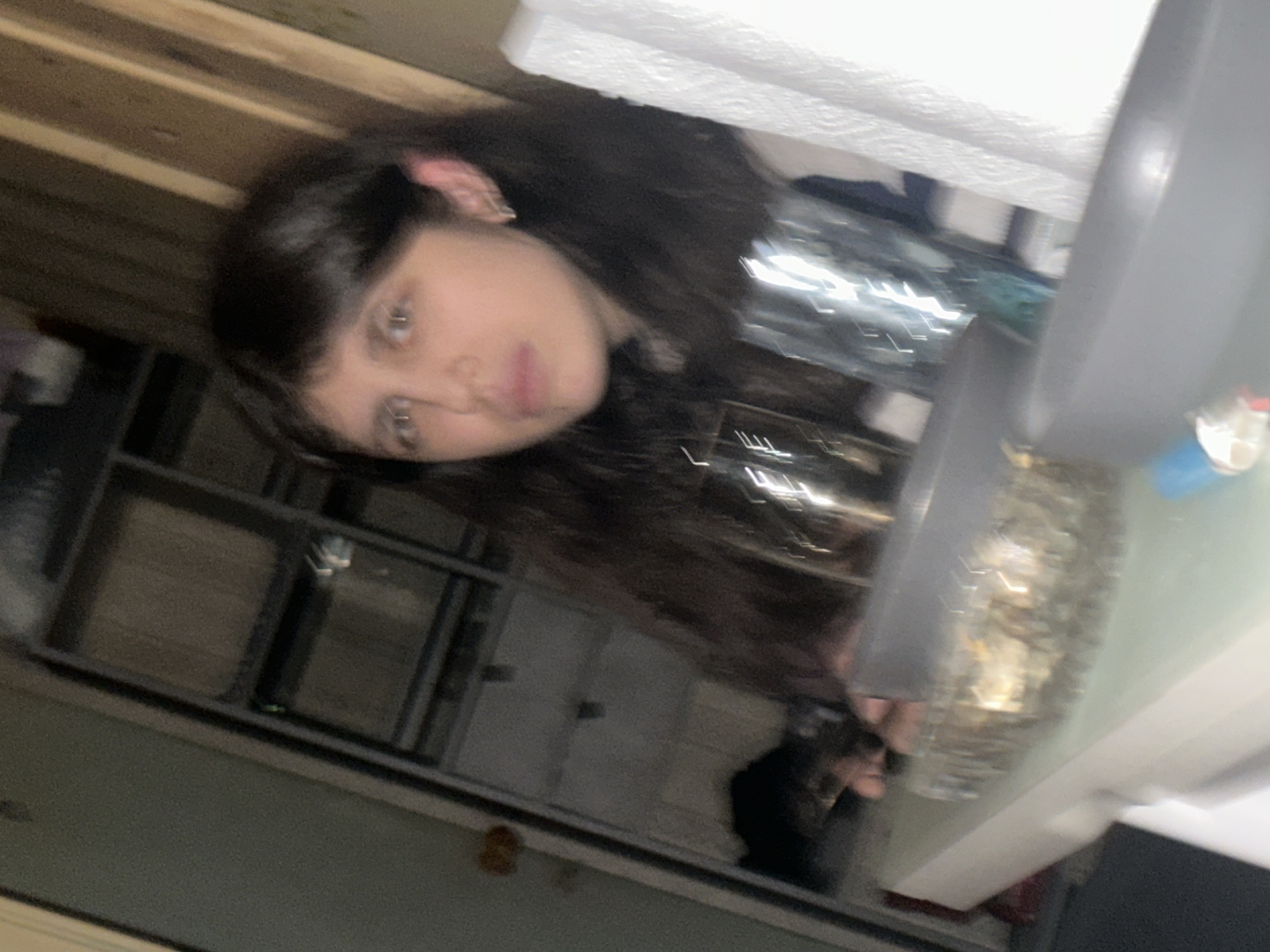 A person with long dark hair and fair skin is looking at the camera. The person is near a kitchen sink with a faucet and some dish soap. A wire rack sits above a counter. The background is blurry, and appears to be an interior space. The image contains reflections that make it difficult to distinguish the objects' forms. There is not enough information to infer that the location is Moscow, Russia.