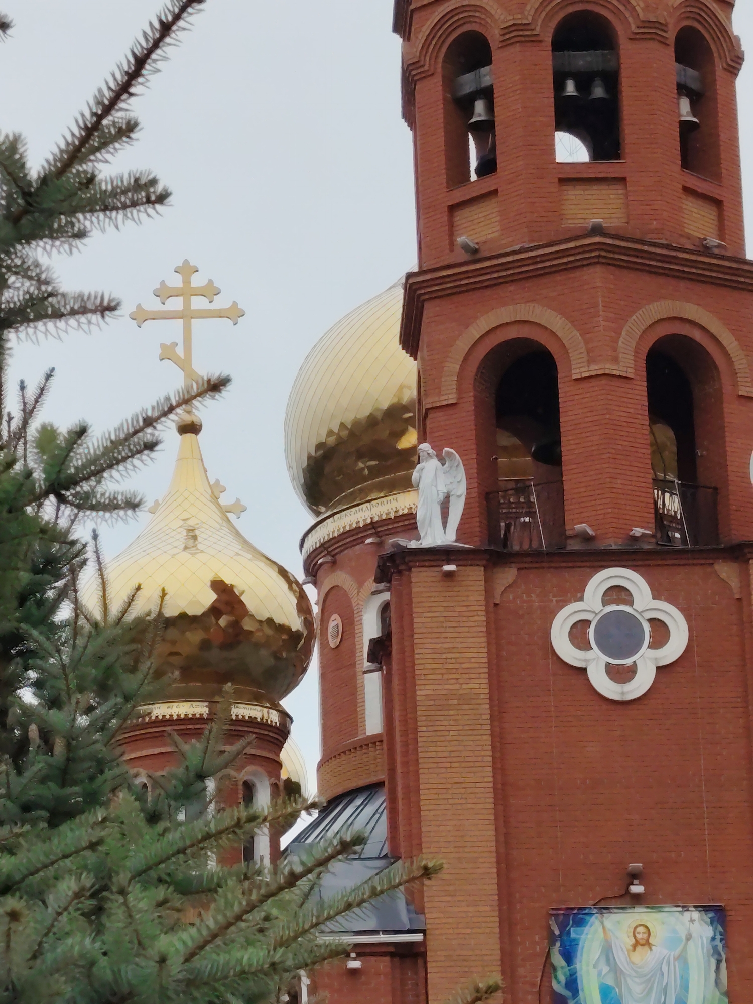 The image displays an Orthodox Christian church in Chaikovsky, Russia, showcasing its distinct red brick architecture and golden domes under an overcast sky. In the foreground, green coniferous tree branches partially frame the view.
Two prominent golden onion domes reflect the diffused light; the smaller dome on the left is crowned with a three-barred Orthodox cross. Around the base of the larger dome, Cyrillic text is visible, which appears to be a dedication, possibly reading "ХРАМ СВ. БЛГ. КН. АЛЕКСАНДРОВСКИЙ" (Church of St. Blessed Prince Alexander).
To the right, a red brick bell tower rises, featuring arched openings that reveal several dark bells within. A white statue of an angel with wings stands on a ledge below the bell openings. Further down the wall, a decorative white quatrefoil motif is integrated into the brickwork. Below this, a vibrant rectangular icon or mural depicting Jesus Christ, possibly illustrating the Resurrection or Transfiguration with a luminous aura, is affixed to the wall.
The scene focuses on the architectural and symbolic elements of the church, with no apparent activity taking place. The construction materials include red brick, gold-plated domes, and white stone or plaster for the statue and trim, all set against a muted sky.