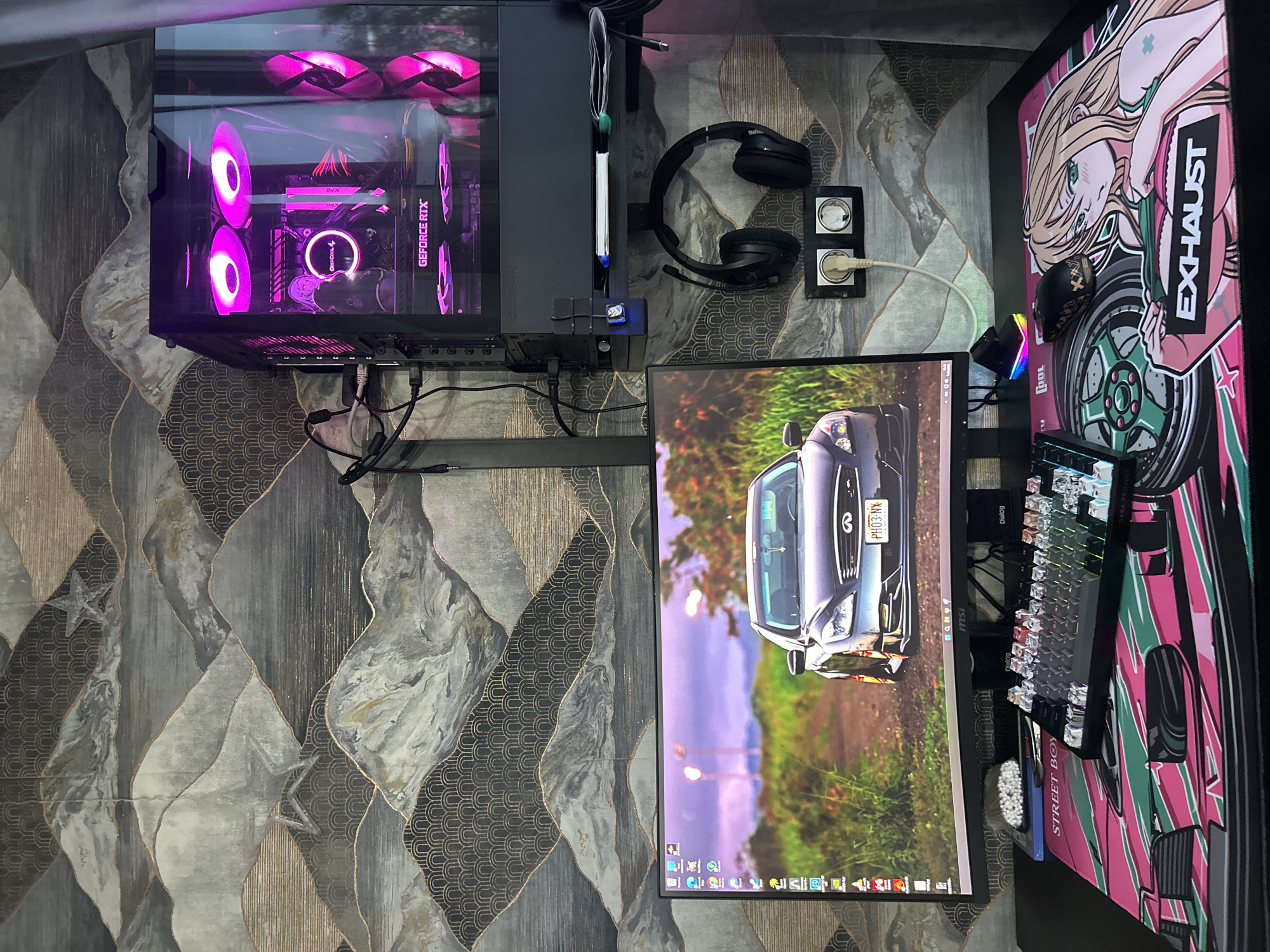 The image shows a computer setup on a desk. A computer tower with illuminated fans and a graphics card is on the left. To the right of the tower are black headphones and a power outlet. A computer monitor displays an image of a silver Infiniti car. A keyboard and mouse are placed on a large anime-themed mousepad. The desk sits on a patterned surface. The location in Chaikovsky, Russia is not ascertainable from the image.