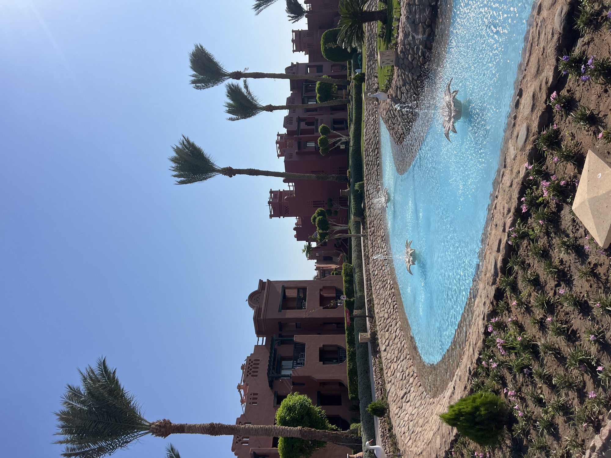The image depicts a resort area in Sharm El Sheikh, Egypt, characterized by its arid climate and distinctive architecture. The scene features a winding, bright blue water channel with decorative fountains, surrounded by stone pathways and meticulously landscaped gardens. Several palm trees of varying heights are scattered throughout the scene, adding to the tropical ambiance.
In the background, a row of multi-story buildings with red-hued facades dominates the landscape. These buildings exhibit a traditional architectural style, with arched windows, balconies, and intricate rooftop details, suggestive of a resort or hotel complex. Lush green hedges line the perimeter of the buildings, contrasting with the dry earth and scattered vegetation.
A white bird statue, resembling a crane or stork, is prominently placed near the water channel. The sky is a clear, cloudless blue, indicating a sunny day. The overall impression is one of a well-maintained and aesthetically pleasing resort environment, designed to offer a tranquil and picturesque experience for visitors.