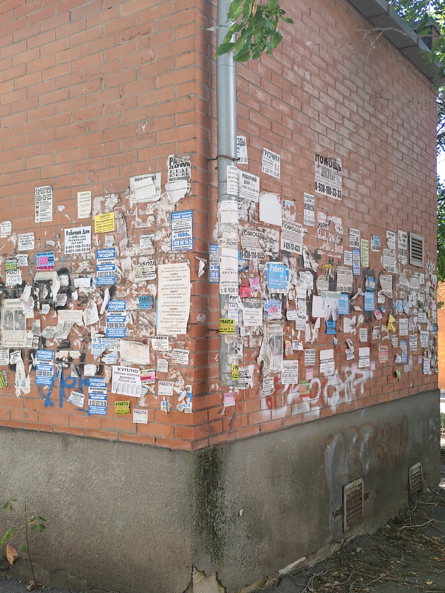The image shows a brick building corner with numerous advertisements and flyers pasted on the wall. The building is located in Rostov-on-Don, Russia, as indicated by the presence of Russian text on the flyers. The flyers advertise various services such as job opportunities, home repairs, and personal services. Some flyers are tattered and partially obscured, suggesting they have been on the wall for some time. The building appears to be old, with a worn concrete base and some graffiti visible at the bottom. The scene is outdoors, under natural light, with a tree branch visible at the top of the image. There are no people visible in the image.