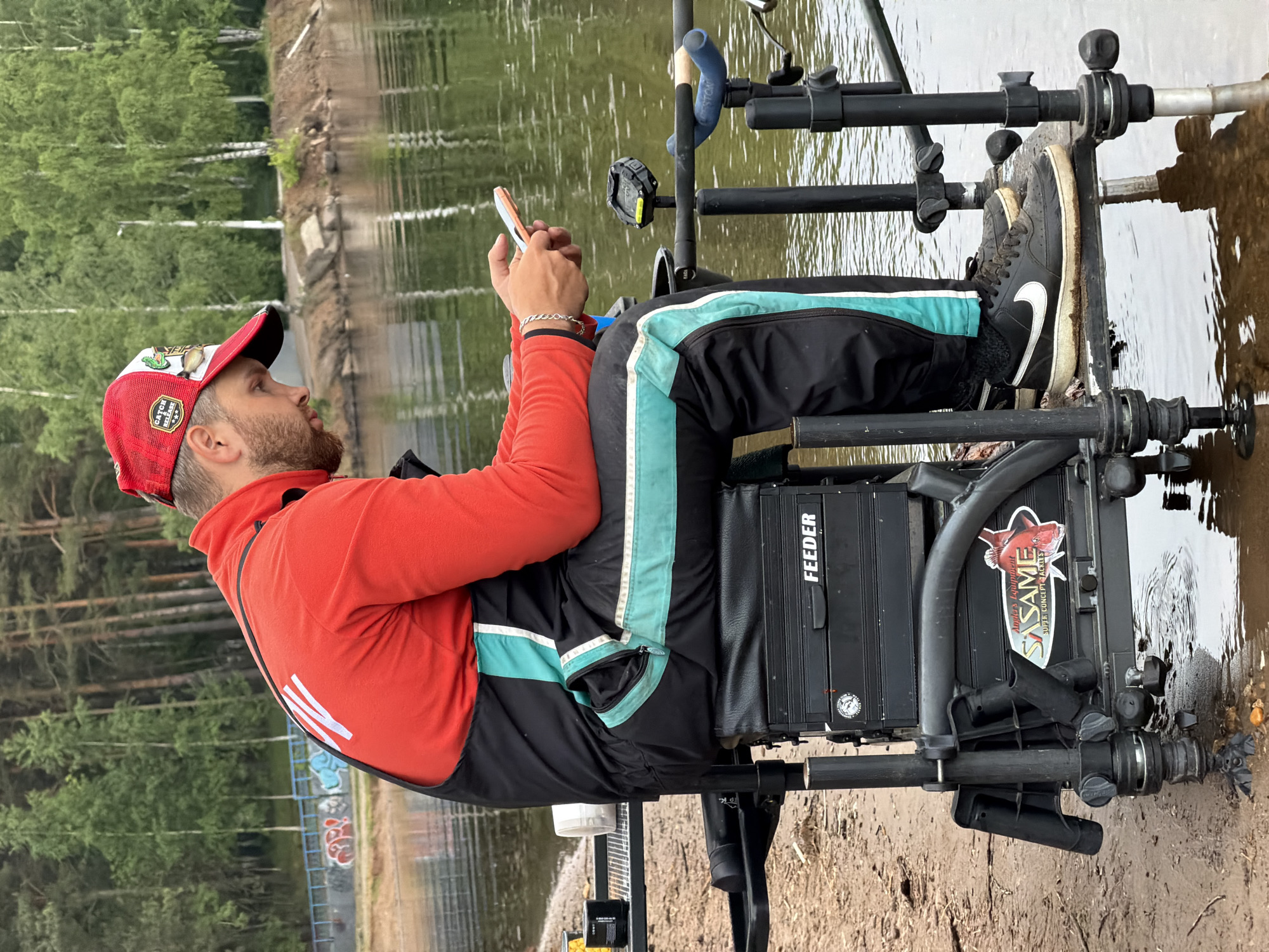 A man sits on a fishing platform on the shore of a body of water, holding a mobile phone. He wears a red and white cap, a red sweatshirt, and black trousers with a light blue stripe. The platform is elevated, and the man’s feet, clad in black sneakers with white swooshes, rest on the platform, partially submerged in the water. Fishing rod supports and other fishing equipment are attached to the platform. The word "FEEDER" is visible on a container. The background shows a shoreline with trees, and the water reflects the sky and vegetation. The image was taken on the shore of the Kama River near Chaikovsky, Perm Krai, Russia.