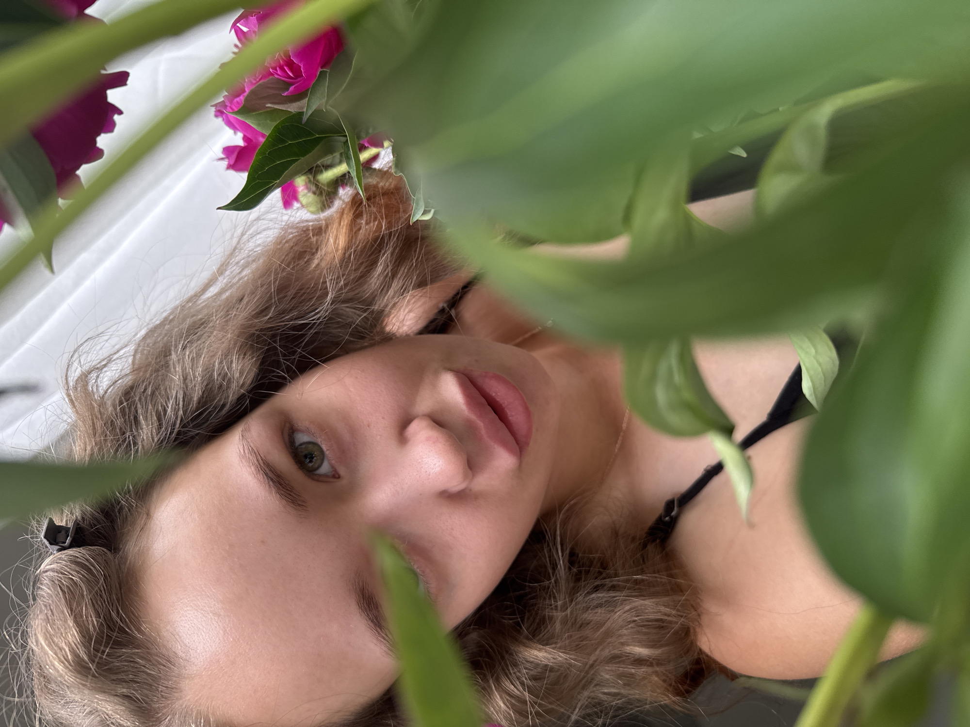 A young woman with light brown curly hair, secured with a hair clip, is positioned in the foreground of the image, with lush green leaves and pink peonies obscuring portions of her face and body. She is looking directly at the camera with her lips slightly parted in a playful pout. Her eyes are a striking green. She is wearing a black garment, possibly a bikini top or dress, with thin straps visible. The background is a light-colored, textured surface, potentially a wall or curtain, with diffused lighting. The overall impression is an intimate, candid portrait taken indoors or in a sheltered outdoor space, possibly a domestic setting in Chaikovsky, Russia, given the presence of flowers which are common in Russian gardens.