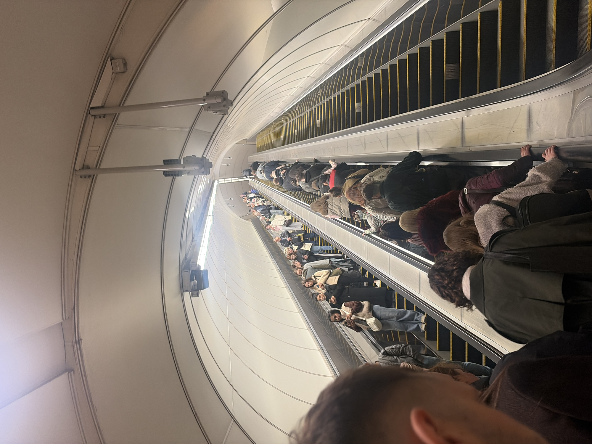The image shows a crowded escalator in what appears to be a metro station in Saint Petersburg, Russia. Many people are riding the escalator, facing downwards. The station has a curved, white tiled ceiling. The escalator's steps are black with yellow edges. People are wearing various outerwear like jackets and coats, with some also wearing hats and carrying bags.