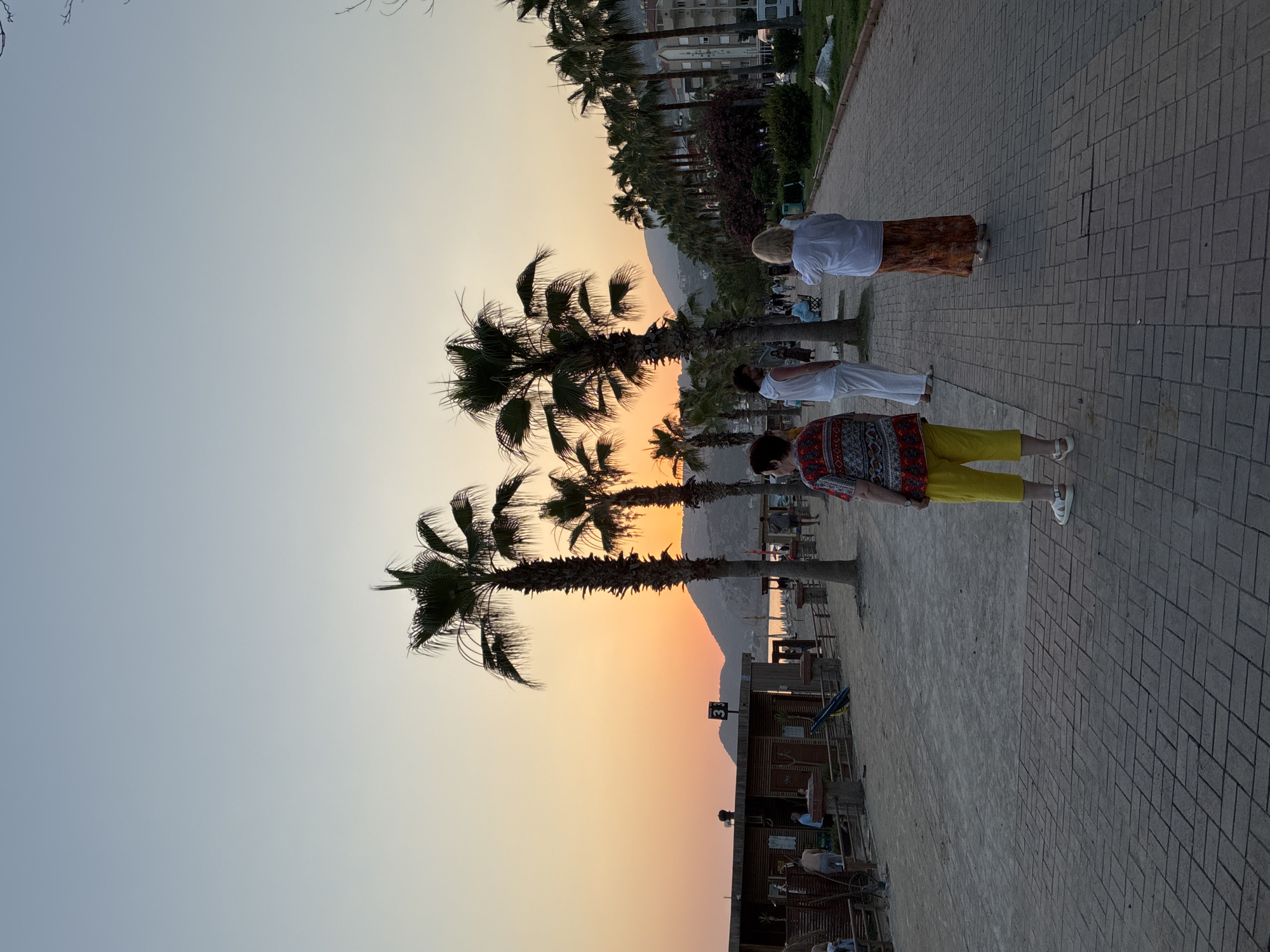 The image depicts a scenic view of a palm-lined walkway beside a beach in Alanya, Turkey, during sunset. Several people are visible on the walkway, casually strolling or standing. In the foreground, a person wearing a patterned shirt and yellow pants is walking away from the camera. Further up the path, another individual in a white top and an orange patterned skirt is seen from the back. A third person, also in a white top and white pants, is walking towards the camera. The background features a row of palm trees, with more in the distance, framing a view of buildings and a mountain range. The sky is a gradient of soft blues and oranges, indicating the sunset. The ground is paved with gray brickwork. The overall atmosphere appears relaxed and peaceful, consistent with a warm evening at a tourist destination. There is a sign with the number "3" visible on a wooden structure in the middle ground.