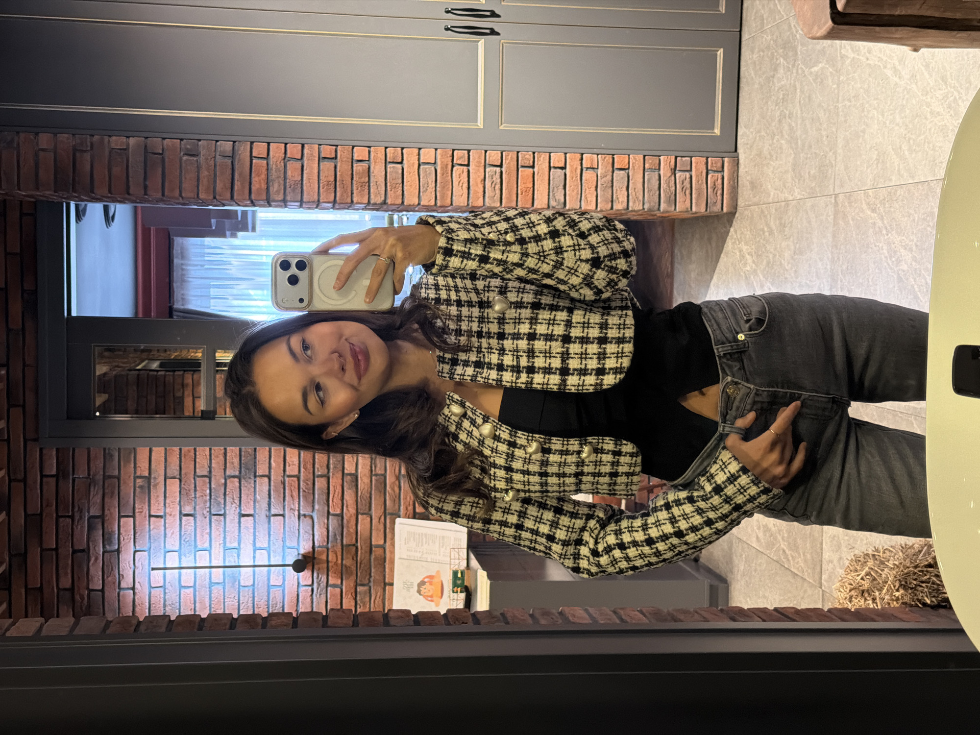 A young woman is taking a selfie in front of a mirror. She is wearing a black top, grey jeans, and a black and white tweed jacket. The reflection shows a room with brick walls, grey cabinets, and tile floors. A window with sheer curtains is visible in the background. A small straw decorative object is present on the floor. The location appears to be in Krasnoyarsk, Russia.