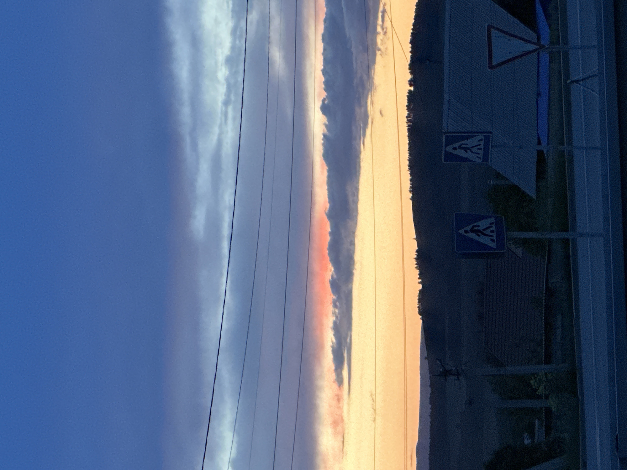 The image depicts a twilight scene near a road in Uchaly, Russia. The sky is a gradient of blue to orange, with a strip of pink-tinged clouds. Power lines run across the sky. On the right, there's a roadside area with a pedestrian crossing sign and a triangular warning sign. The background includes some trees and buildings, obscured by shadow.