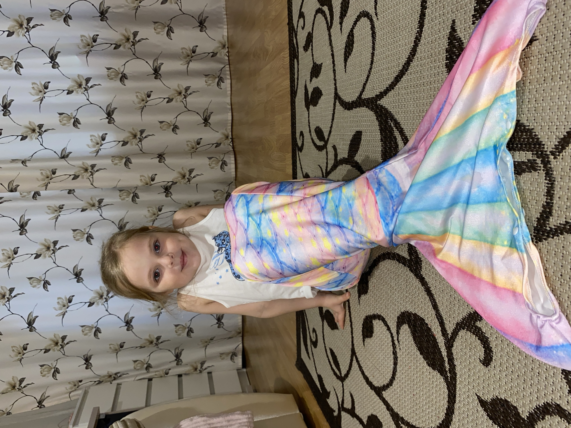 A young child is depicted in an indoor setting, rotated 90 degrees clockwise from an upright perspective. The child, likely a girl with blonde hair and light-colored eyes, is wearing a vibrant, multicolored mermaid tail costume, predominantly featuring pastel pink, blue, yellow, and white in a tie-dye or rainbow pattern, complete with a fin. Underneath the costume, she wears a white tank top with a blue design of a stylized animal head (possibly a leopard or cheetah) surrounded by stars on the front; some partially visible text, possibly "LA" or "CA", is beneath the design.
She is lying on a light-colored rug with an intricate dark brown or black floral/vine pattern. A strip of light brown wooden flooring is visible next to the rug, separating it from white or off-white curtains adorned with a repeating pattern of light brown floral branches and leaves. A glimpse of white furniture is visible in the bottom left corner. The child is looking directly at the camera with a slight smile and an engaging expression, appearing happy and content while playing or posing in her costume. The overall scene suggests a cozy, domestic environment.