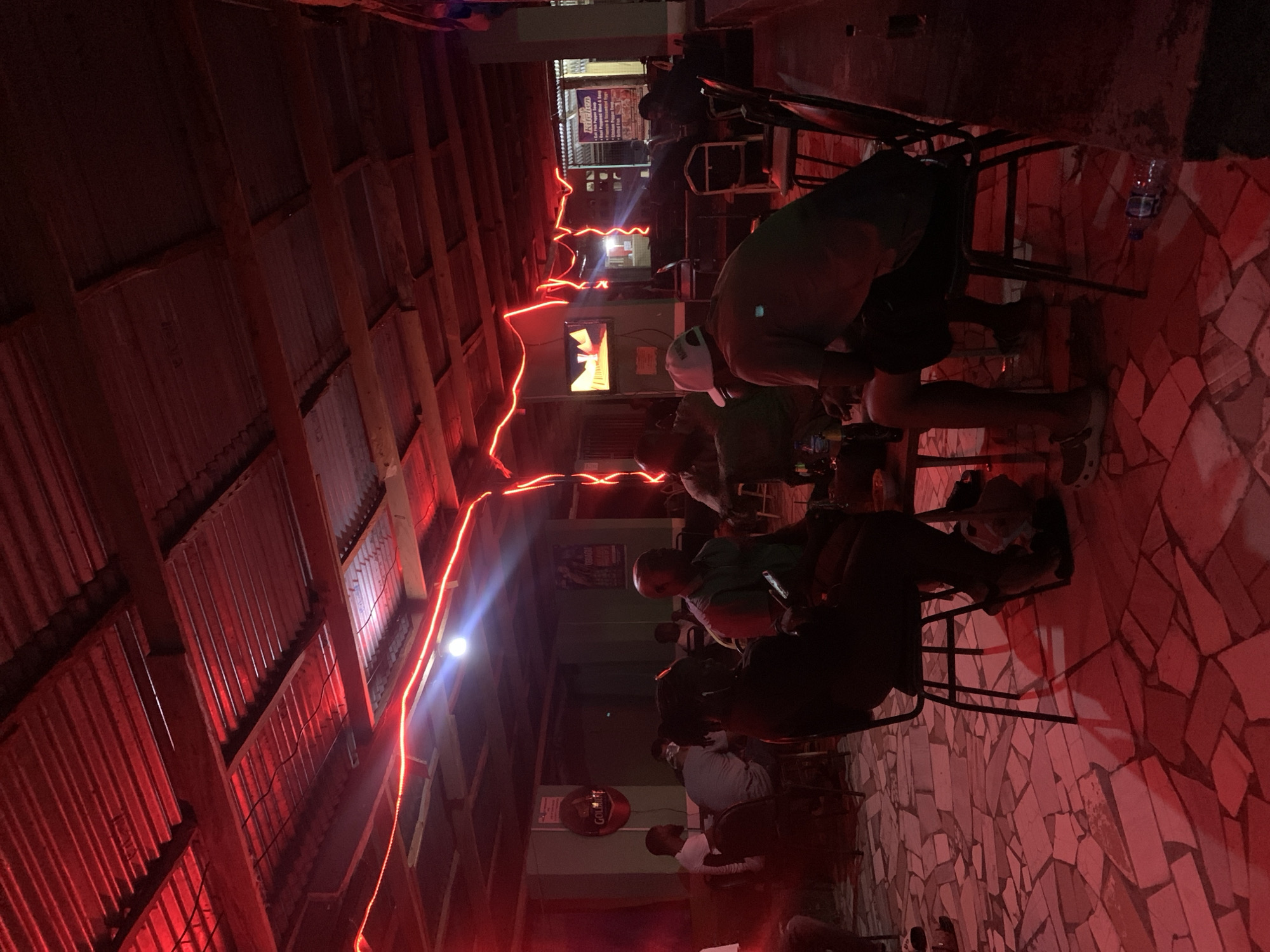 The image depicts a nighttime scene inside a bar in New Stadium, Nigeria. The bar has a tin roof illuminated by a string of red lights. Several people are seated on metal-framed chairs. A television screen is visible on one wall. The floor is tiled with red and white mosaic pieces. A water bottle rests on the floor near a chair. Several people are interacting in the scene.