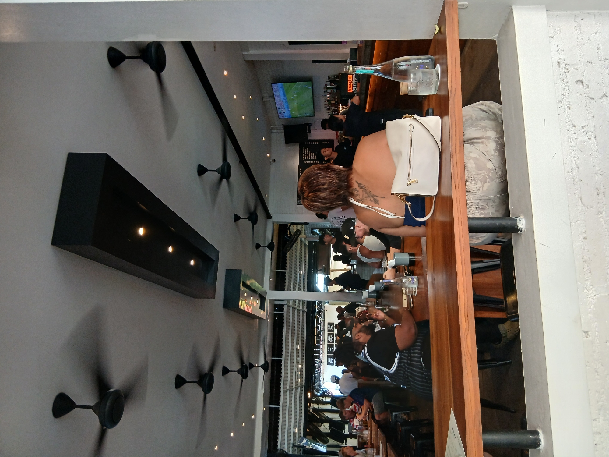 The image depicts the interior of a bar or restaurant in North Kuta, Indonesia. Multiple ceiling fans are visible, some in motion. The scene shows a bar area with patrons seated at the bar and at tables. A television screen is mounted on the wall behind the bar, displaying a sports game. Bottles of alcohol are stocked behind the bar. In the foreground, a person is seated at a wooden table, with a handbag and a glass of water on the table. The overall ambiance appears casual and social.