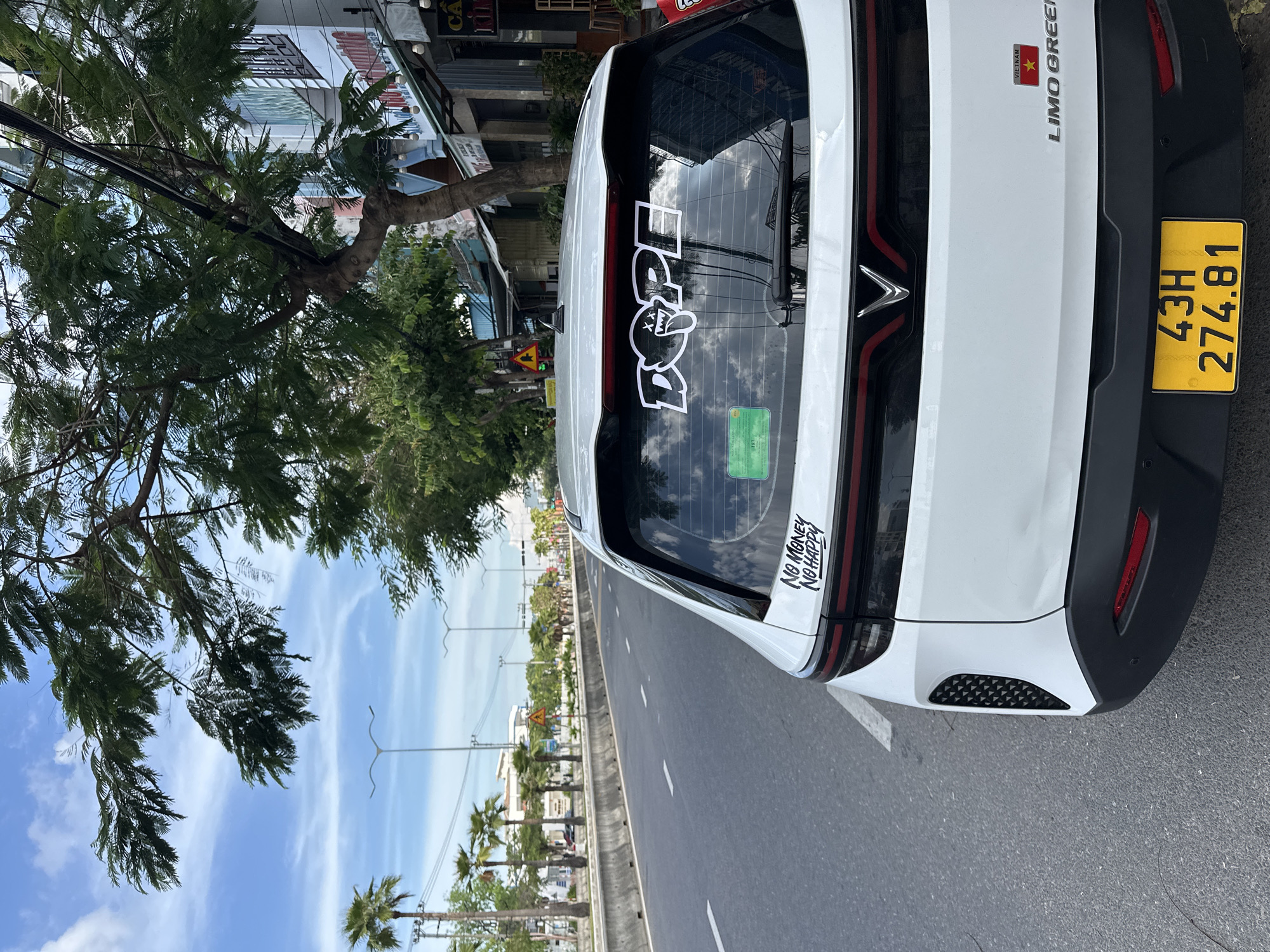 The image captures a white VinFast VF e34 electric car parked on the side of a street in Đà Nẵng, Vietnam. The car's rear is prominently featured, displaying two stickers on the back window: "DOPE" in large, stylized lettering with a cartoon character's face, and a smaller sticker that reads "NO MONEY NO HAPPY". A Vietnamese license plate, yellow with black characters, is visible on the lower rear bumper, reading "43H 274.81". The car is positioned on an asphalt road with white lane markings. To the left, a concrete divider separates the road from a sidewalk lined with palm trees and lampposts. In the background, buildings with awnings and signage are visible, suggesting a commercial area. The sky is partly cloudy with a blue hue. The overall scene depicts a street view in a Vietnamese city on a clear day, with the focus on the parked car and its embellishments.