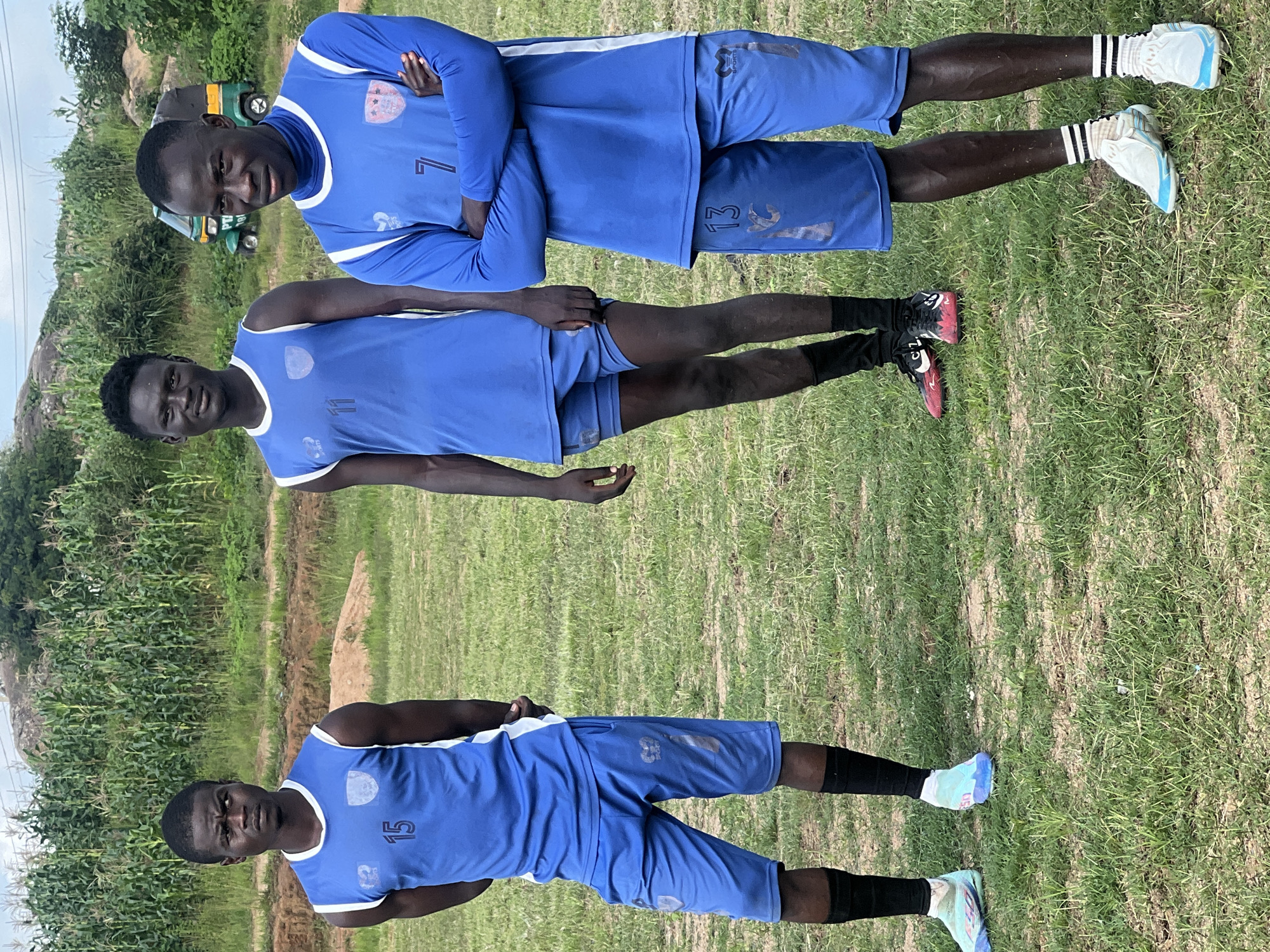 Three young men in matching blue soccer uniforms stand on a grassy field in Jos, Nigeria. The man on the left wears a sleeveless jersey with the number 15 and blue shorts. The man in the center wears a sleeveless jersey with the number 11 and blue shorts. The man on the right wears a long-sleeved jersey with the number 7 and blue shorts, with his arms crossed. They are all wearing athletic shoes. In the background, a field of green crops and a green and yellow three-wheeled vehicle are visible. The sky is overcast.