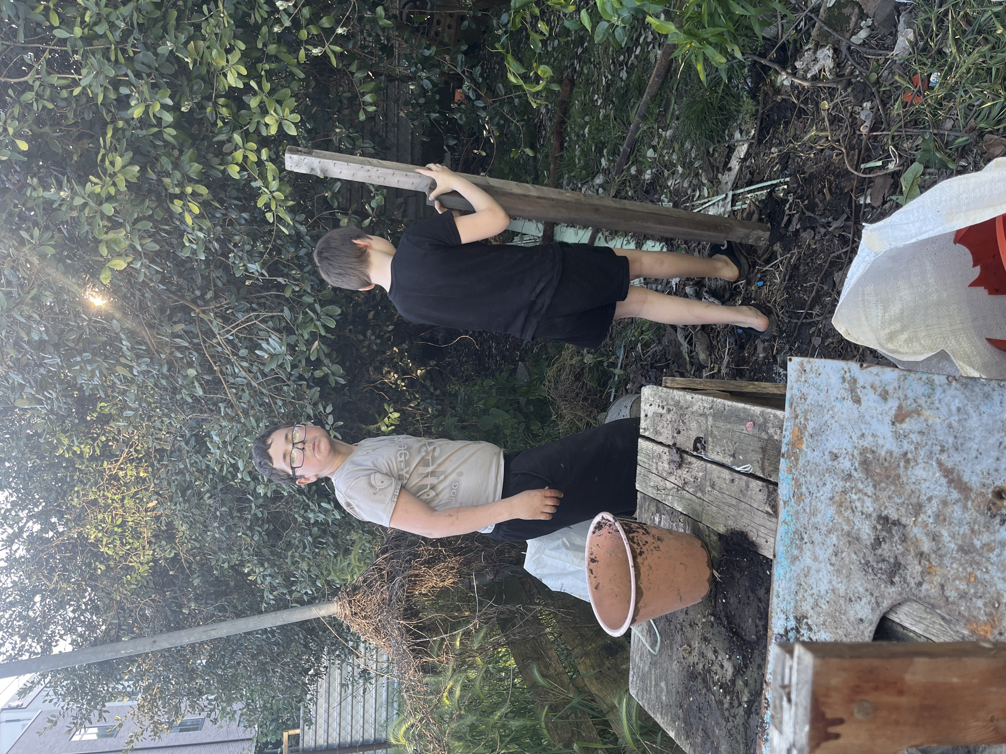Two boys are in a cluttered outdoor area with trees and plants. One boy wears glasses, a light-colored t-shirt, and dark pants, while the other wears a black t-shirt and black shorts. The boy in black carries a wooden beam. Various objects such as a bucket, wooden planks, and a metal surface are visible. The location is not definitively discernible as Vesyoloye, Russia, based solely on the visual information provided.