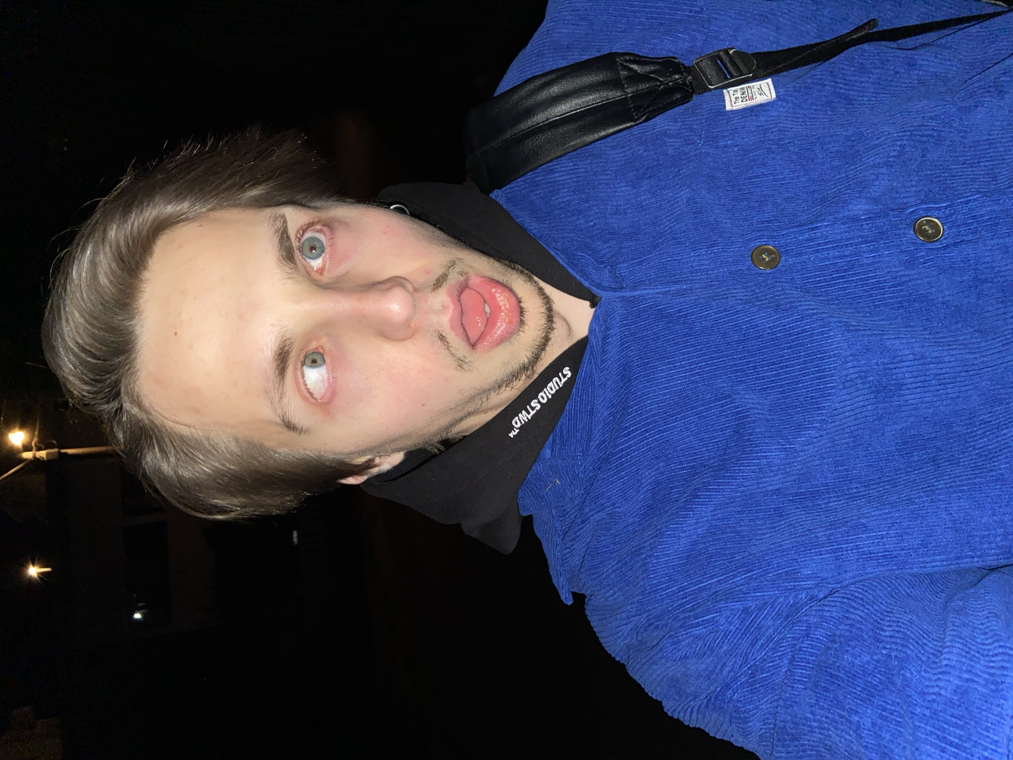 A disoriented young man is pictured in an informal outdoor setting in what appears to be Šušanj, Montenegro. The man, who has fair skin, light eyes, and light-brown hair, is wearing a blue corduroy jacket over a black hooded sweatshirt. The hood of the sweatshirt is visible around his neck. A black strap, potentially from a bag, crosses his shoulder. The man has a short beard and mustache and is sticking out his tongue. The background is dark, but a street lamp and window can be seen on the left side of the frame. The image is rotated counterclockwise.