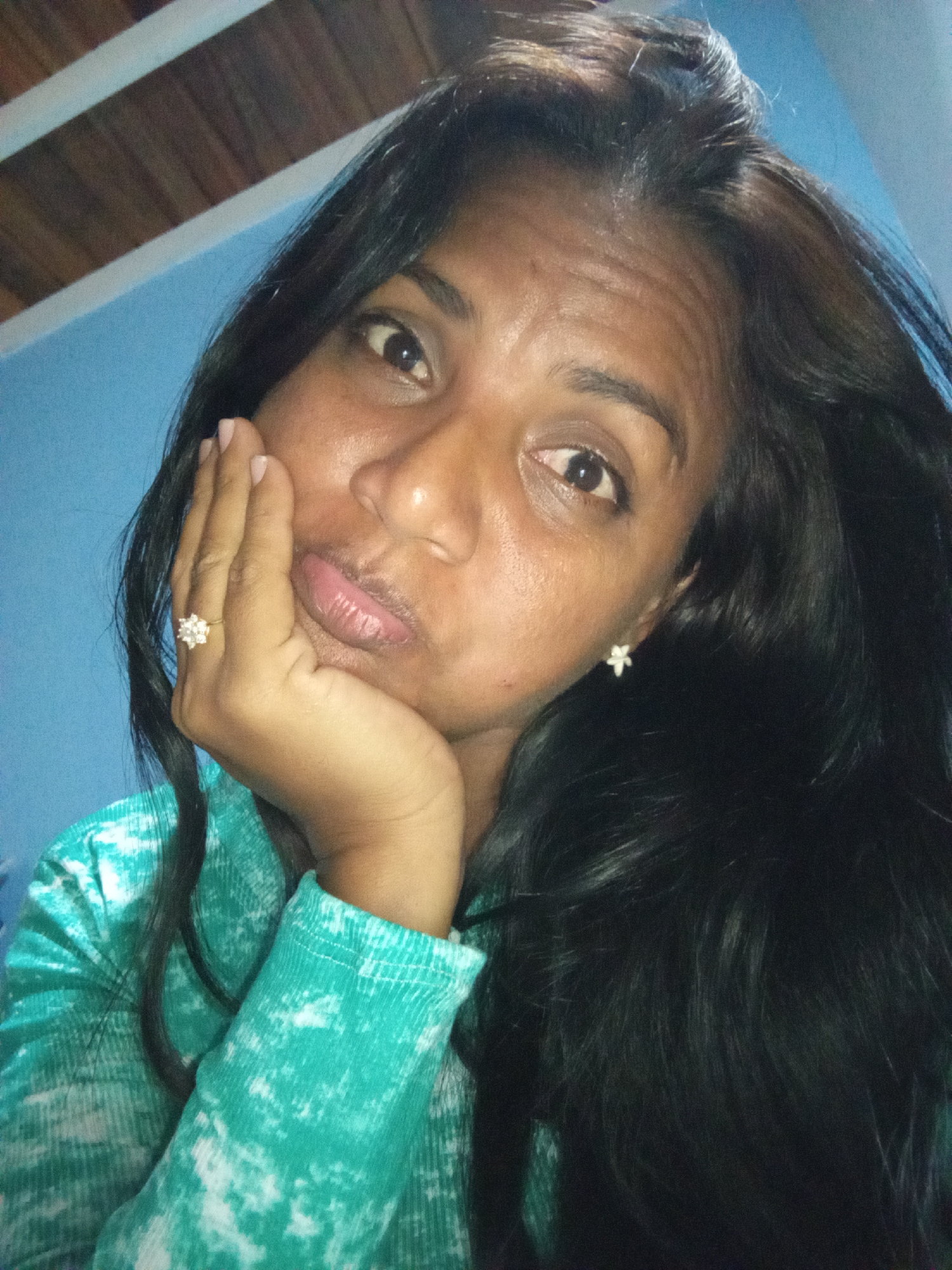 A close-up selfie shows a woman resting her chin on her hand, her right cheek supported by her palm. She has dark, long hair that falls around her shoulders and chest. Her eyes are open and directed towards the camera, and her lips are slightly pursed. She is wearing a teal-colored long-sleeved shirt with a tie-dye pattern of white splotches. A ring with multiple small stones is visible on her right hand, and a small, white, star-shaped earring is on her left ear. The background consists of a light blue wall and a wooden ceiling with white beams. The lighting is dim.