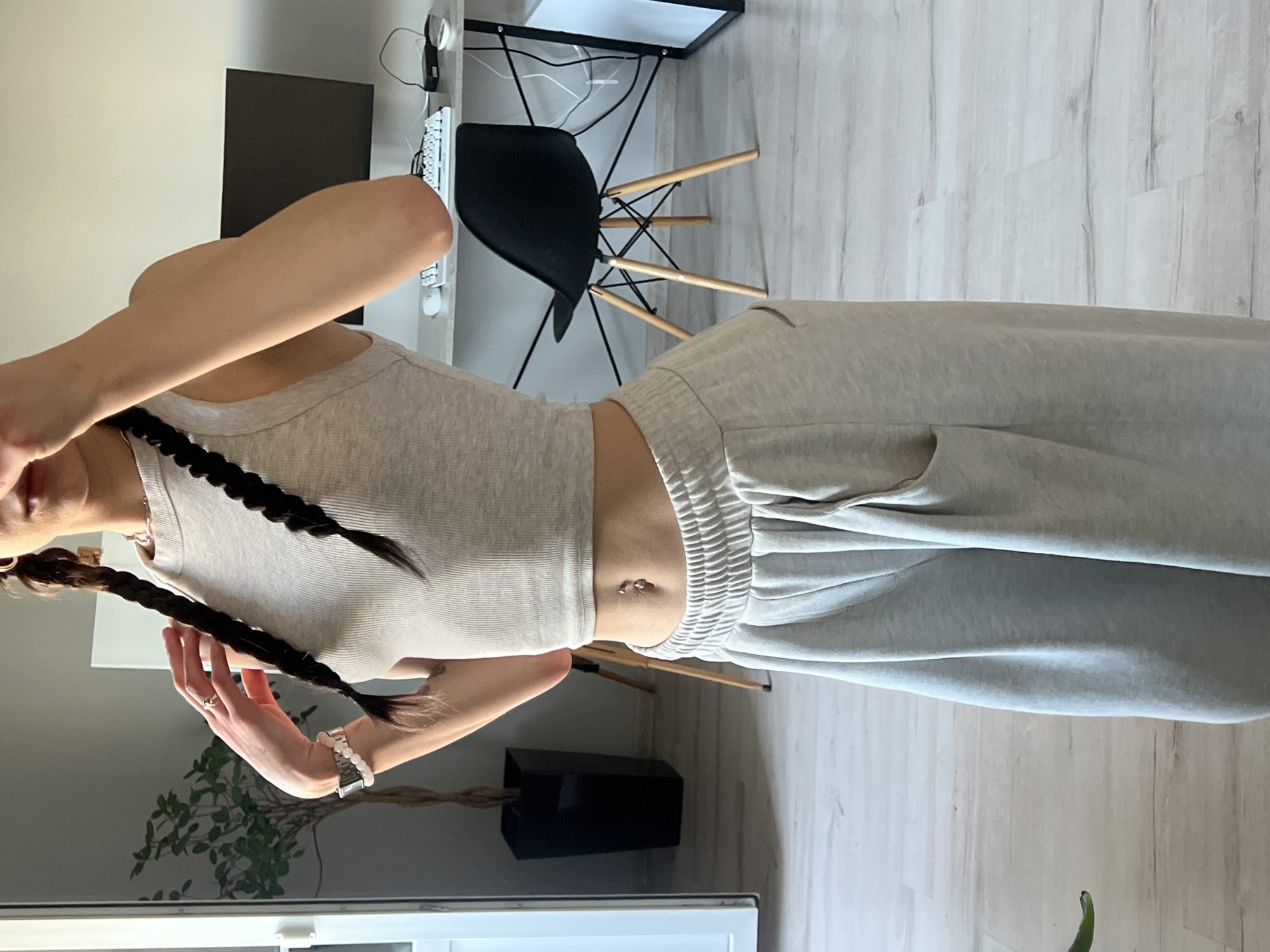 The image shows a woman standing in an indoor setting, likely a room with a desk and a chair. The woman is wearing a beige ribbed tank top and light gray sweatpants. She has her hair in two braids and is wearing a watch and a ring on her left hand. She also has a belly button piercing. In the background, there is a desk with a computer monitor, keyboard, and mouse, as well as a black chair with wooden legs. There is also a tall plant in a black planter. The floor is made of light-colored wood planks. There are no identifiable landmarks or features to suggest the location is in Ternopil, Ukraine.
