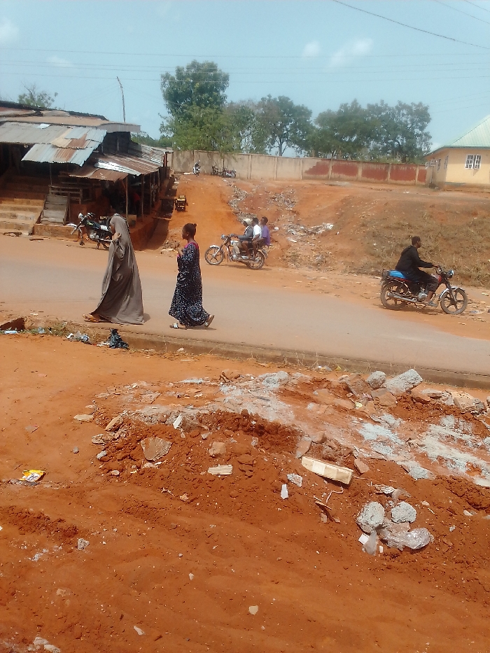The image shows a street scene in Auchi, Nigeria (latitude: 7.05, longitude: 6.263). There are three people walking on the left side of the street. One is wearing a grey abaya and headscarf, one is wearing a long black and white dress, and one is wearing a blue and white dress. There is a group of three people on a motorcycle in the background. A man is riding a motorbike on the right side of the street. The street is paved, and there is a dirt shoulder on the left side of the street. There is a dirt pile with debris in the foreground, and there are buildings and trees in the background.