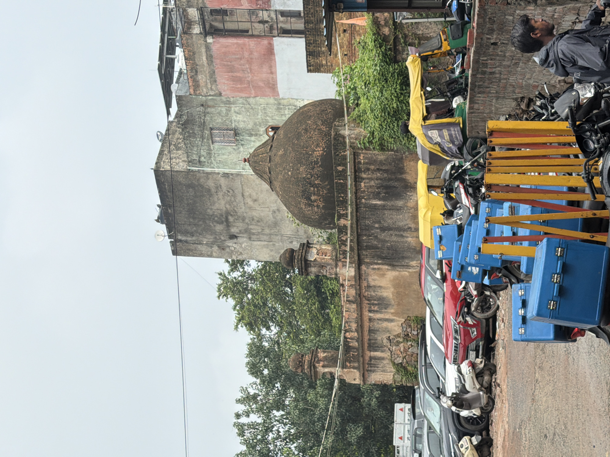 In an outdoor setting, a parked red SUV is visible behind a white scooter, facing left. In front of the SUV, a white scooter is parked. Further left, a white truck is visible. A large dome-shaped structure with a mottled dark brown surface stands behind a weathered stone wall. The dome is topped with a small finial. The wall has decorative stone pillars at its ends. Behind the dome and wall, several trees with lush green foliage are present. In the foreground, several blue storage containers are stacked. A person in a dark jacket is visible on the right side of the image, looking to the left. The scene appears to be in a city, possibly Delhi, India, with vehicles and buildings suggesting an urban environment.