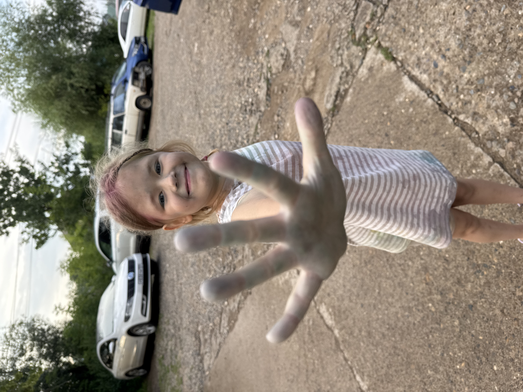 A young girl with pink-streaked hair and a smudged face smiles at the camera, holding up her hand, which appears to have been in paint. She is wearing a striped dress and stands on a cracked concrete surface. In the background, several cars are parked. The scene is outdoors with trees and a cloudy sky visible. This image was taken in the city of Chaikovsky, Russia.