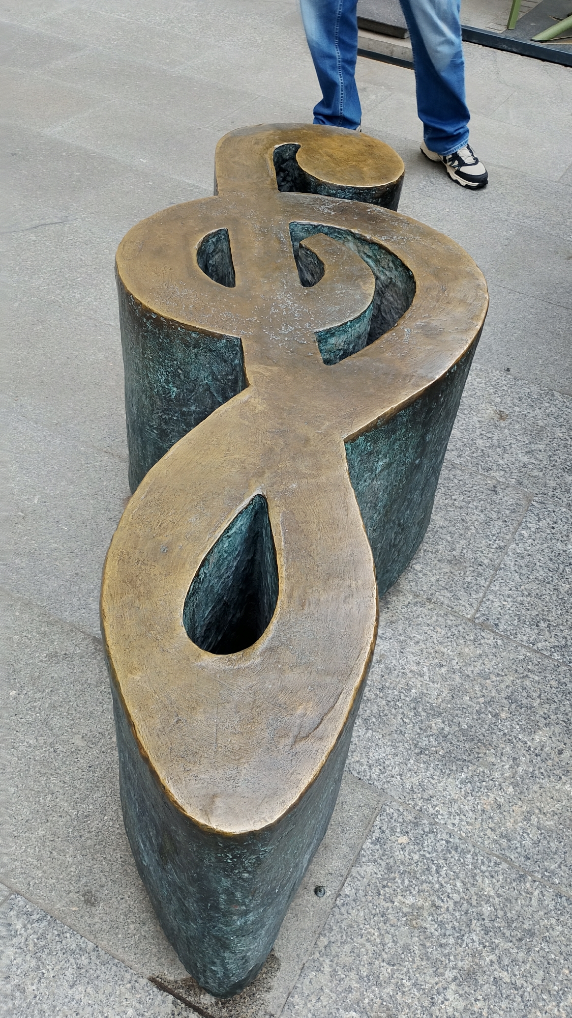 A bronze-colored treble clef sculpture sits on a gray granite tiled surface. A pair of legs and feet belonging to an individual wearing jeans and black-and-white sneakers are seen in the background, with the person seemingly standing near the sculpture. The sculpture itself has a greenish-blue oxidized patina on its sides, with the top surfaces being a lighter bronze color. The location is in Łódź, Poland, specifically along Piotrkowska Street.