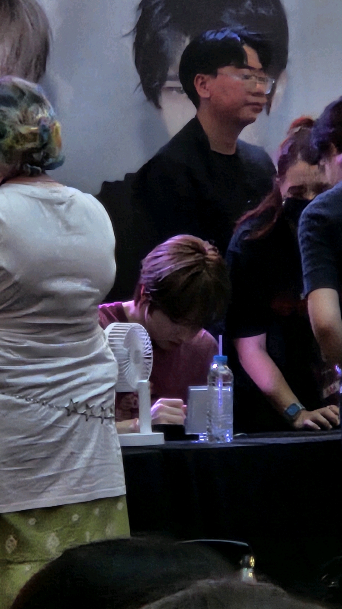 The image shows a group of people at an indoor event, possibly a fan meet or signing, in Barcelona, Spain. In the foreground, a person wearing a white top and a green patterned skirt is partially visible from the back, with colorful hair. To their right, a person with short brown hair wearing a maroon shirt is leaning over a table, appearing to sign something. A small white fan is placed on the table in front of them. A clear plastic bottle with a blue straw is also on the table. Behind this person, a woman with dark hair is wearing a black top. Further back, a man with glasses and dark hair is looking to the side. The background features a large graphic or poster with stylized images of people's faces. The overall lighting suggests an indoor venue with artificial lighting.