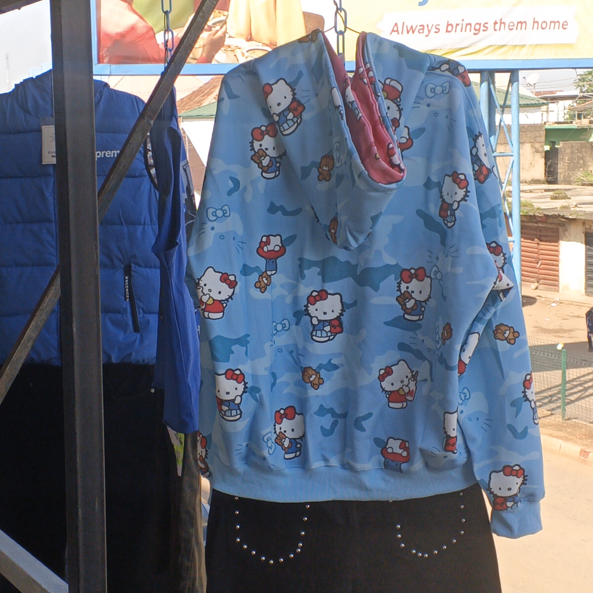 This image captures a street scene in Bukuru, Nigeria, featuring clothing items displayed for sale. In the foreground, a blue puffer jacket with a tag bearing the word "prem" is visible on the left. To its right, a light blue hooded sweatshirt adorned with a repeating pattern of Hello Kitty characters and camouflage detailing is displayed prominently. The sweatshirt has a pink lining visible inside the hood. Beneath the sweatshirt, a black skirt with a decorative pattern of silver studs is partially visible. Behind the clothing, a yellow banner with red text reading "Always brings them home" is mounted. The background reveals elements of the urban environment, including buildings constructed from concrete blocks, a metal fence, and a paved street. The overall lighting suggests it is daytime, with the sun casting shadows.
