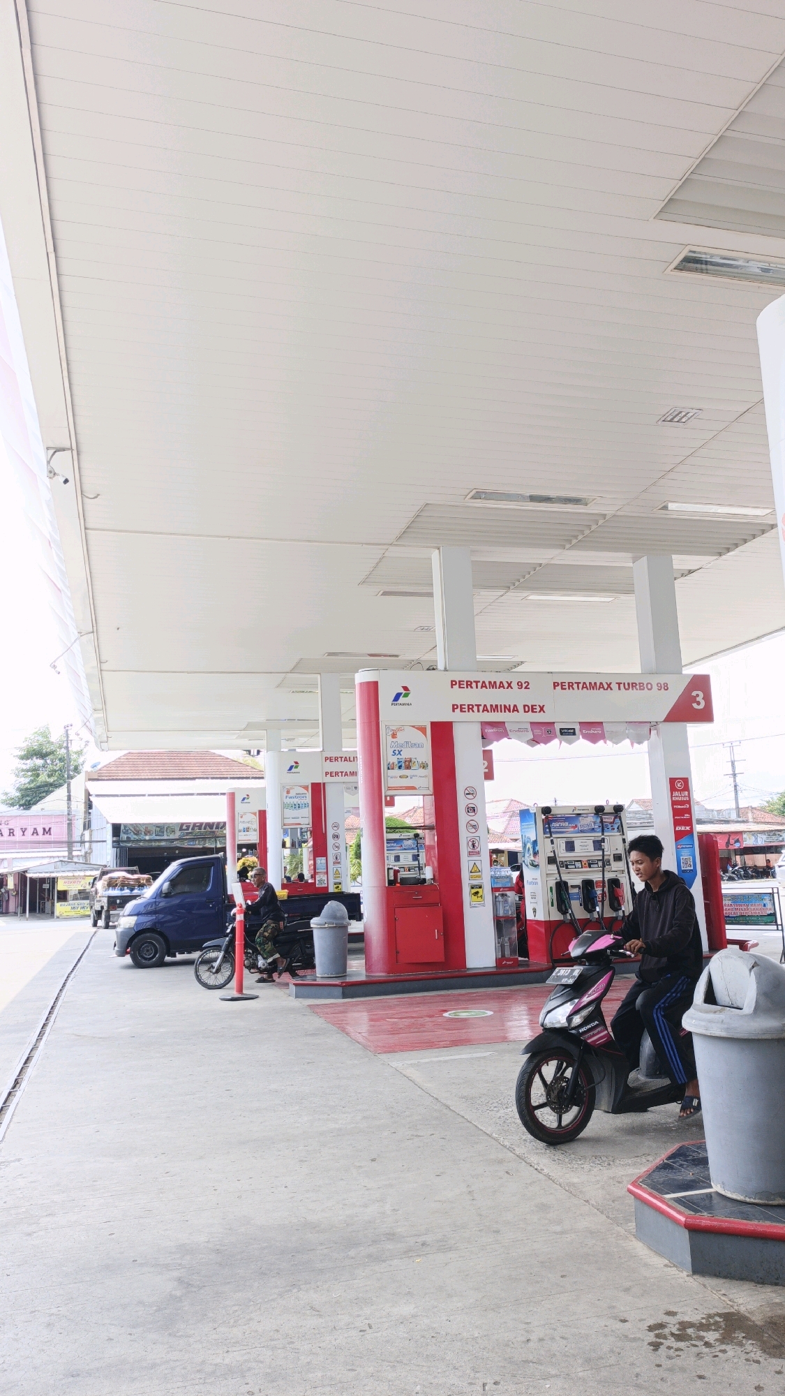 The image depicts a scene at a Pertamina gas station in Desa Wonoharjo, Indonesia. In the foreground, a young man wearing a dark hoodie and striped track pants is seated on a black and pink scooter, facing right. He is looking towards the gas pumps. To his left, a blue pickup truck is parked. Further left, another motorcycle is parked, with a person in camouflage clothing seated on it, facing away from the camera. The gas station features red and white pumps with Pertamax 92 and Pertamax Turbo 98 branding. There are trash cans visible at the station. In the background, buildings and trees are present, suggesting an outdoor, urban or semi-urban environment. The sky is overcast.