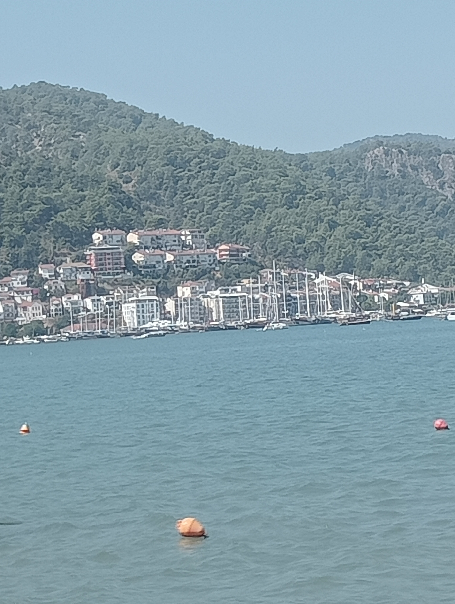 The image shows a panoramic view of a coastal town nestled at the base of a lush, green mountain range. The town, which appears to be Fethiye, Turkey, features a dense collection of buildings with red-tiled roofs cascading down the hillside towards the waterfront. A prominent harbor filled with numerous sailboats, many with their masts erect, lines the shore. The water in the foreground is a calm, blue expanse dotted with a few buoys, one orange and another partially visible red one. The sky is a clear, pale blue. The overall impression is one of a picturesque Mediterranean or Aegean coastal scene.