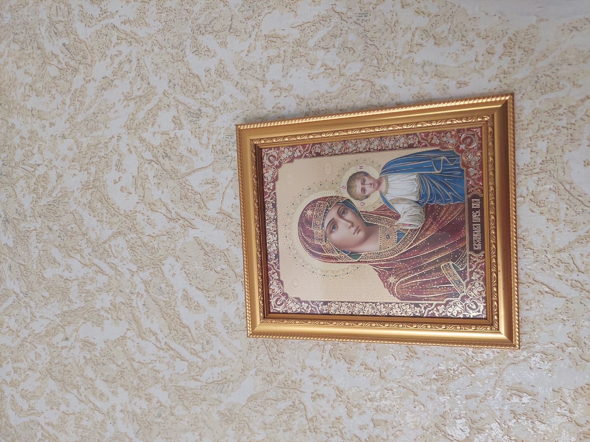 A framed religious icon depicting the Virgin Mary and the infant Jesus is displayed on a textured, beige wall. The icon features the figures with halos and is adorned with intricate patterns and gold accents. The Virgin Mary wears a dark red garment with gold detailing, and the child is dressed in blue and white. Below the image, there is text in a foreign language. The frame is golden and ornate. The background wall has a rough, uneven surface with a marbled or stucco-like finish in light brown and beige tones. The lighting appears to be natural, casting a soft glow on the scene. The icon is positioned slightly off-center within the frame.
