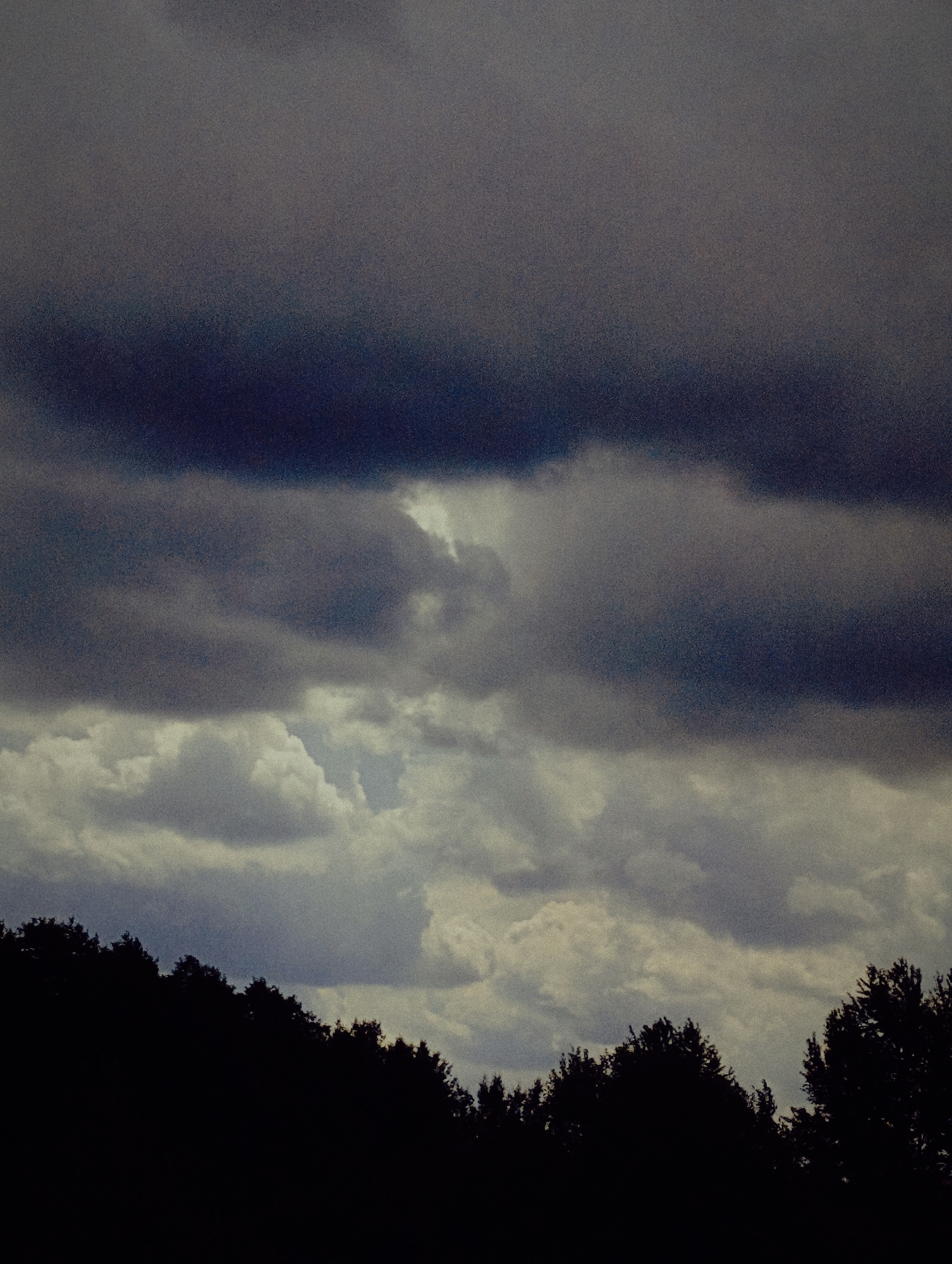 The image shows a silhouette of trees against a dark, cloudy sky. The clouds are dense and appear to be gathering, suggesting an impending storm. The sky is a mix of dark grays and lighter blues, with some areas of brightness indicating breaks in the cloud cover. The trees are dark and appear to be deciduous, with their branches visible against the sky. The scene is captured from a low angle, looking up at the sky. The location is described as Unknown, Unknown, suggesting that the specific city and state are not provided.