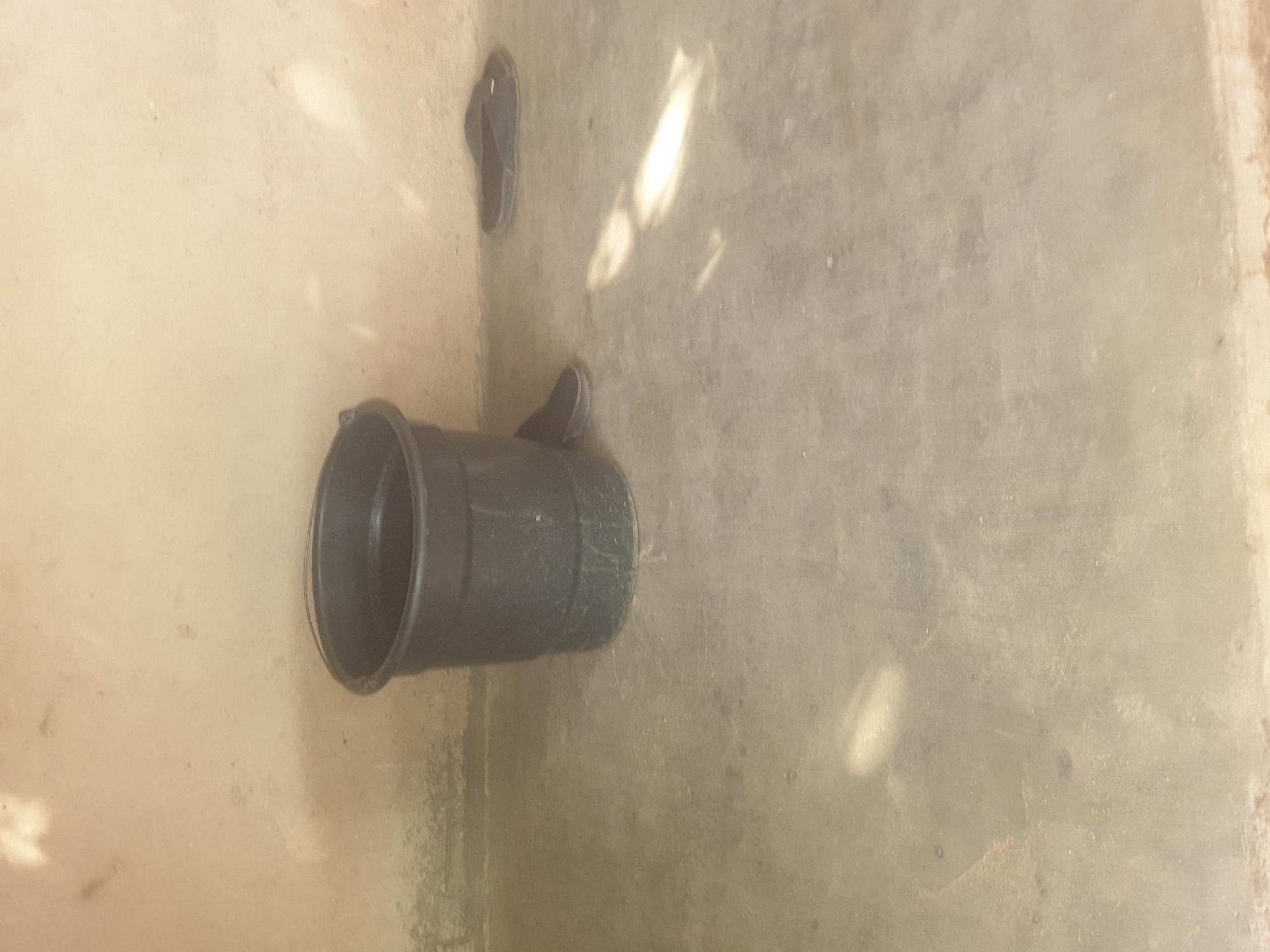 A black plastic bucket sits on a concrete floor against a light-colored wall. To the right of the bucket, a pair of black flip-flops is visible on the floor. The scene is illuminated by natural light, casting shadows and highlights on the floor and wall. The environment appears to be an outdoor or semi-outdoor space, possibly a yard or alleyway. There are no people visible in the image. The location is identified as Jauro Yinu, Nigeria, suggesting this is a street-level view or a residential area.