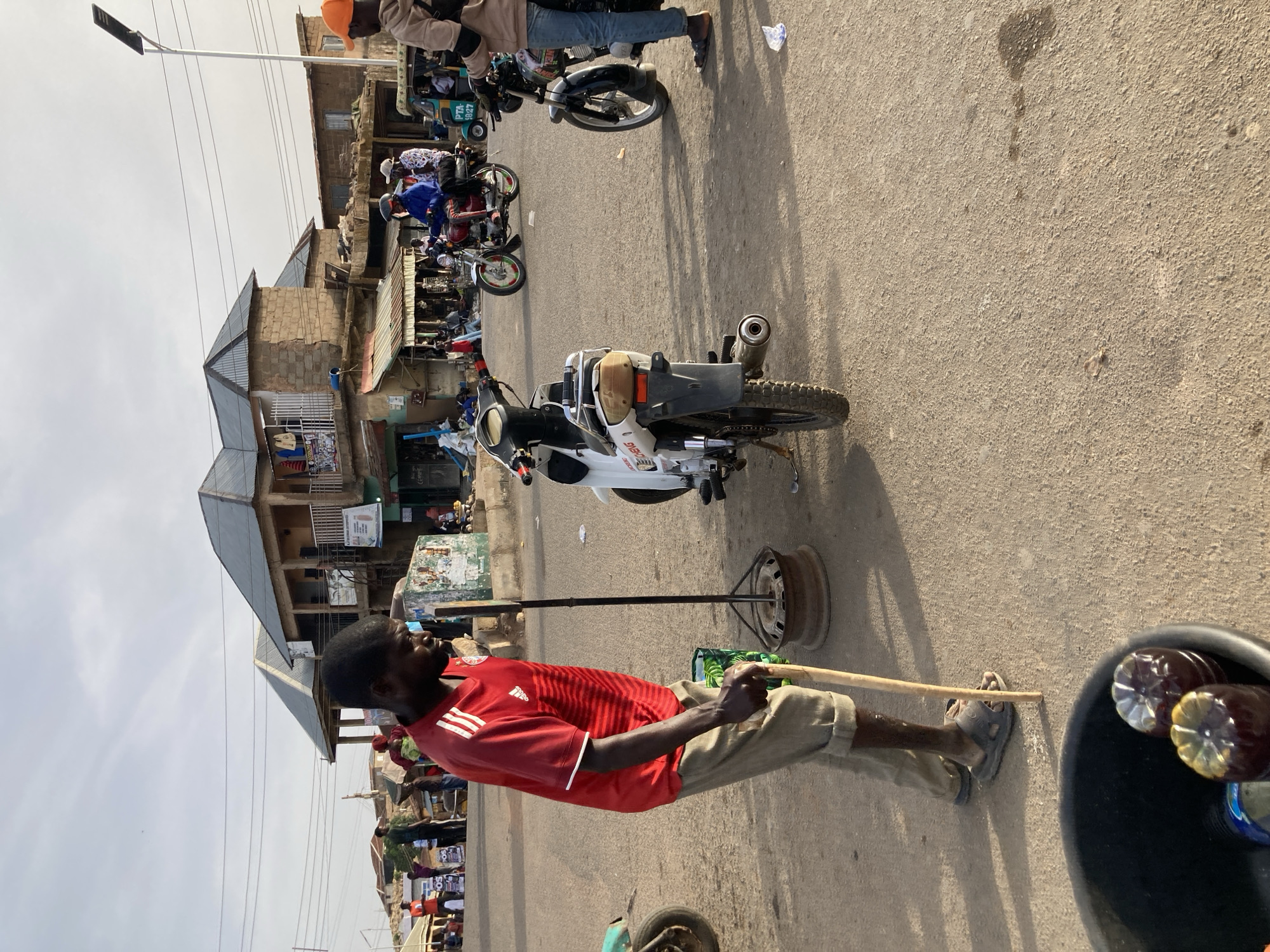 A man in a red and white Adidas shirt and tan shorts is standing on a paved street in Gwong, Nigeria. He is using a long wooden stick to stir something in a metal wheel placed on the ground. A white motorcycle is parked nearby. The street is crowded with people and other motorcycles. Buildings are visible in the background. The sky is overcast. A black bucket with filled water bottles is visible.