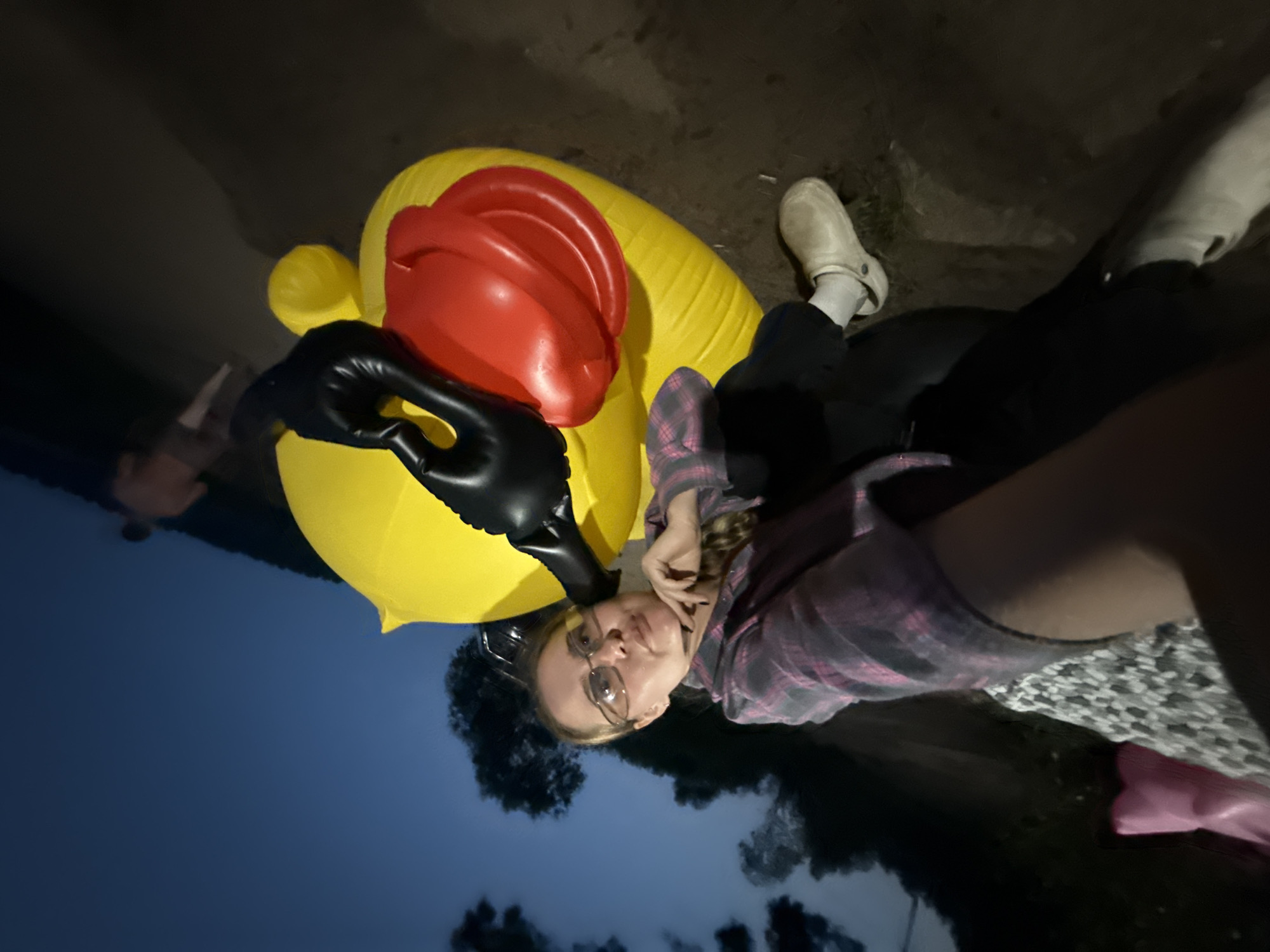 A young Caucasian woman with blonde hair, wearing glasses, a pink and grey plaid shirt, black pants, and white clogs, is photographed in a low-angle, wide shot. She is reclining on an inflatable yellow duck with a red beak and black inflatable wings. Her right hand is resting on her chin, and her head is tilted slightly. The background consists of a dark blue twilight sky with some clouds and trees. To the right, a blurry figure of a person in an orange shirt is visible in the distance. The ground appears to be dirt and gravel. This scene is located on the shore of the Kama River in the city of Chaykovsky, Russia.