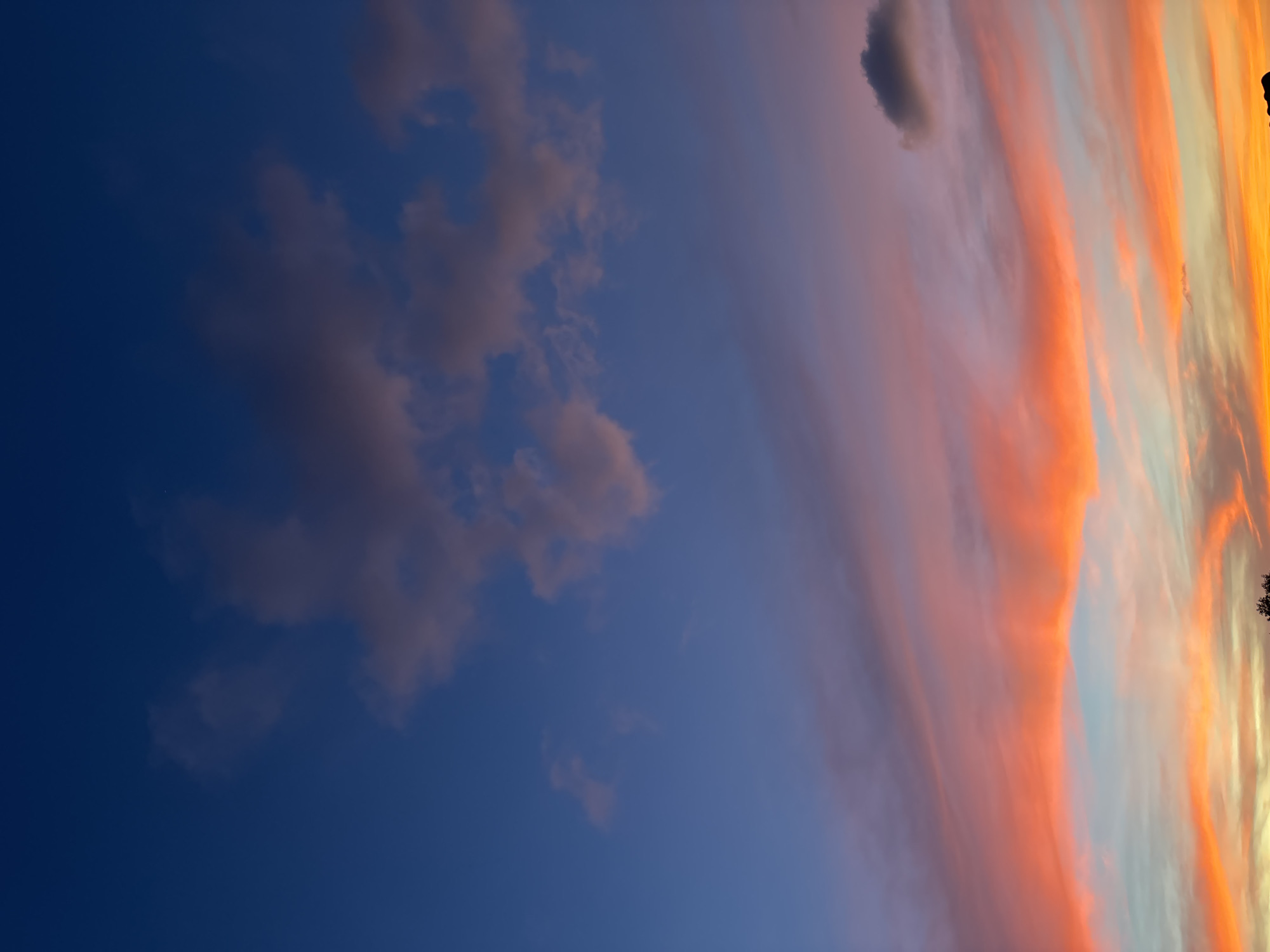 The image captures a sunset sky in บ้านบางรักษ์, Thailand. The sky transitions from a deep blue on the left to vibrant orange and yellow hues on the right. Wispy clouds are present, some tinged with pink and orange, stretching across the scene. A few dark clouds are also visible. In the extreme right corner, silhouettes of trees can be discerned.