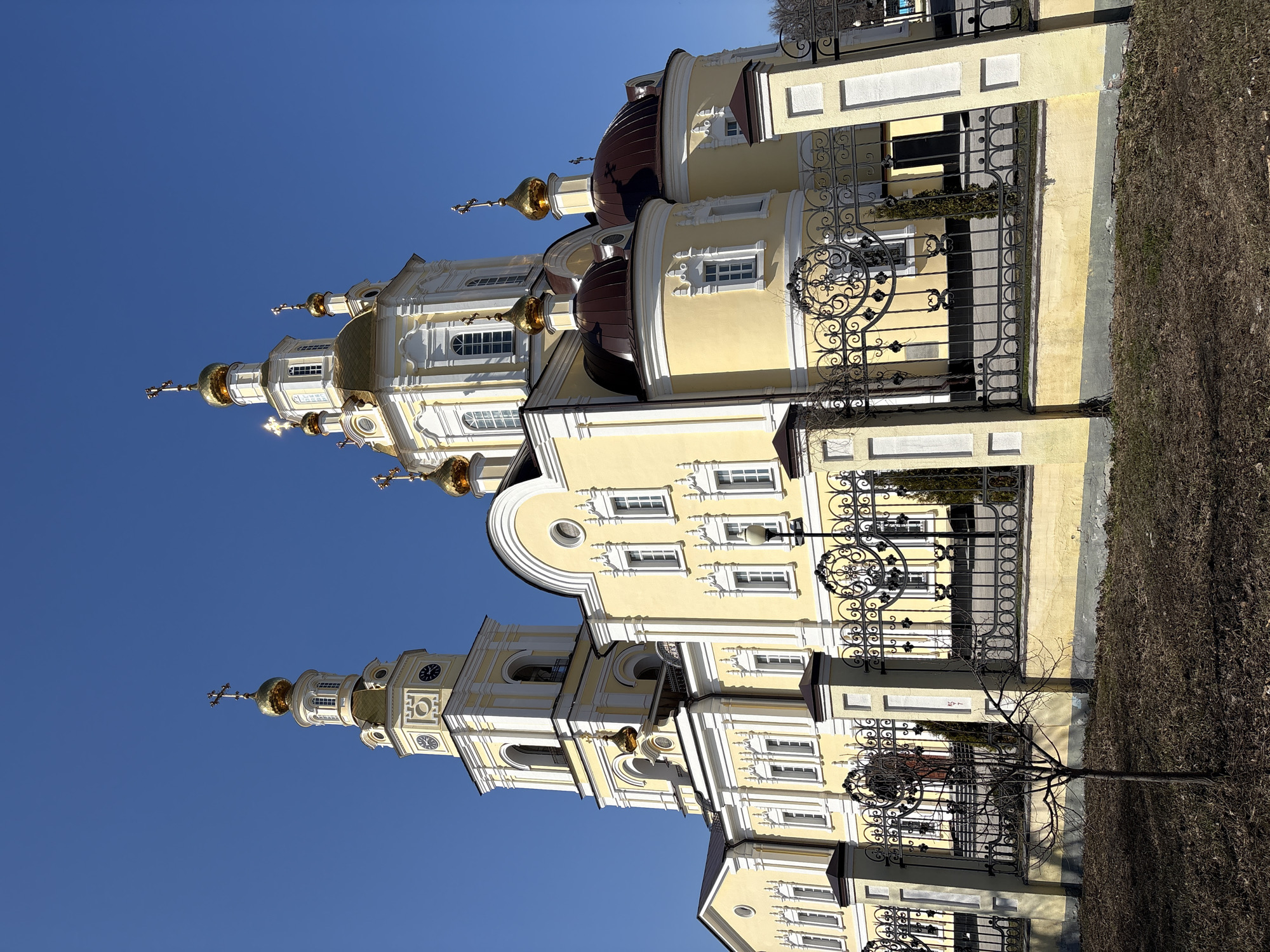 The image shows the Holy Ascension Cathedral in Ulyanovsk, Russia. It is a yellow building with white trim and multiple golden domes topped with crosses. The building has a clock tower. An ornate black wrought iron fence surrounds the property, adjacent to bare shrubs. The sky is clear and blue.