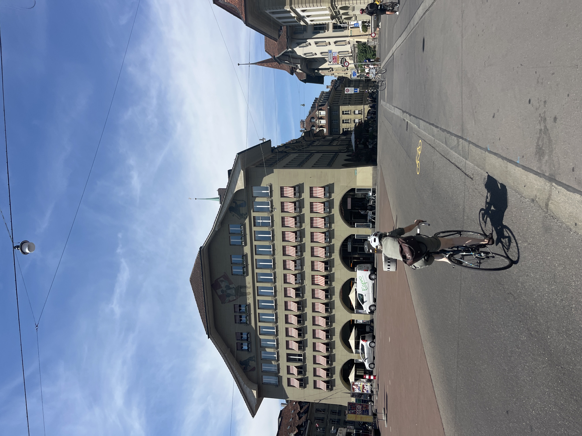 The image depicts a scene in the city of Bern, Switzerland, specifically in the vicinity of Kramgasse or a similar cobblestone square. The foreground shows a person riding a bicycle from behind, facing away from the camera, on an asphalt road. The cyclist is wearing a light-colored helmet, a khaki-colored short-sleeved shirt, and dark shorts, with a brown backpack. The bicycle is dark in color with thin tires.
Dominating the mid-ground is a large, multi-story building with a distinctive green facade. This building features numerous arched entrances on the ground floor, some of which are covered by awnings. Above these arches, the building has multiple rows of windows, many of which are equipped with red and white striped awnings. Faint murals are visible on the upper part of this building. Parked in front of the arches are a white van with "Espro" branding and a silver car.
Further into the background, the street continues, lined with similar historic European-style buildings with tiled roofs and windows. To the right, a building with a pointed spire and reddish-brown roof stands out. The sky is bright blue with scattered white clouds. The overall impression is a sunny day in a European city with a well-preserved architectural style and active pedestrian and bicycle traffic.